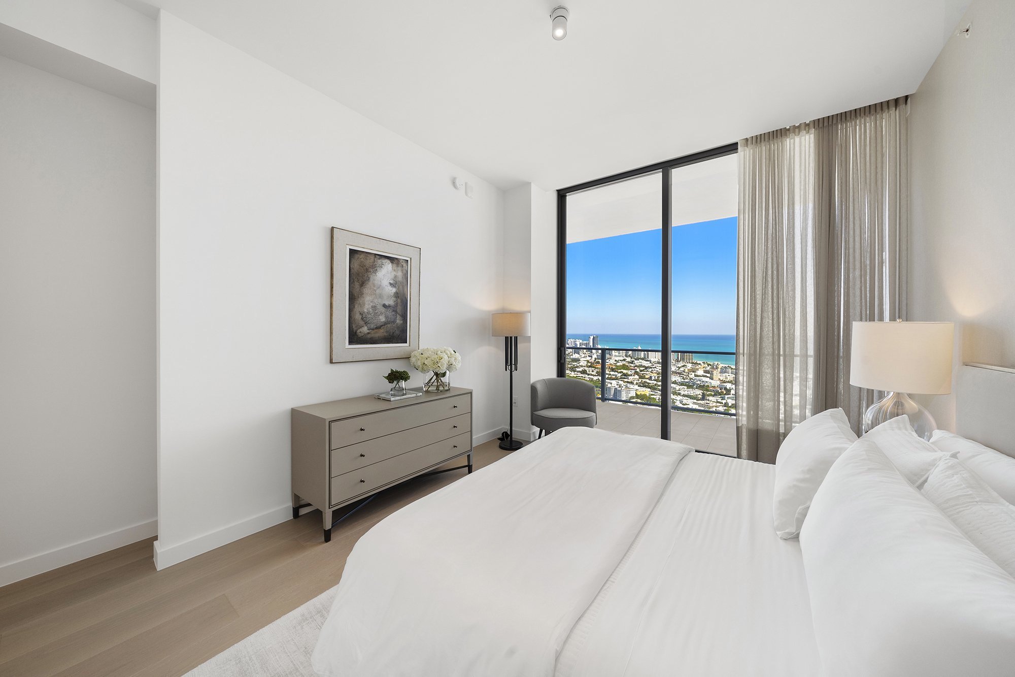 Modern bedroom with white bedding, a dresser, art on the wall, and large windows offering an ocean view.