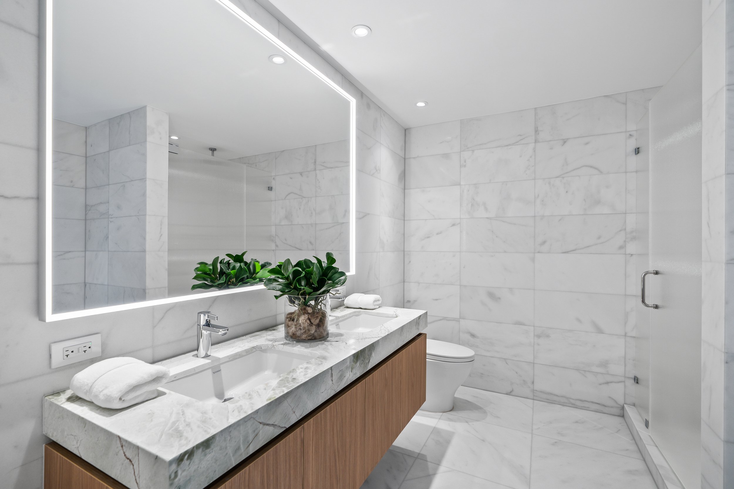 Modern bathroom with marble tiles, large illuminated mirror, vanity with a sink, potted plant, toilet, and glass-door shower.
