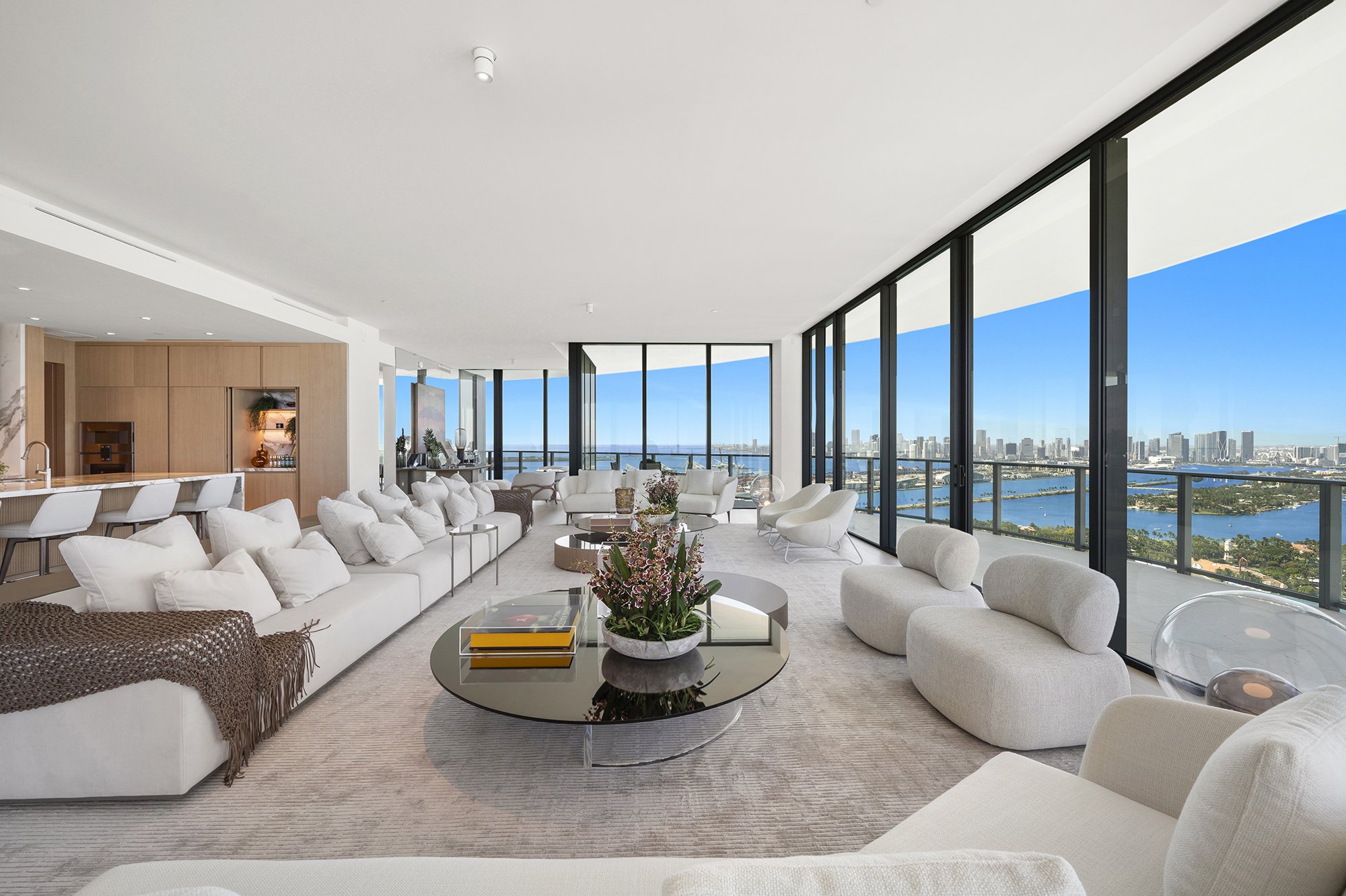 Luxurious living room with large windows offering city and ocean views, featuring modern furniture, a plush sofa, round coffee table with plants, and an open kitchen with bar seating.
