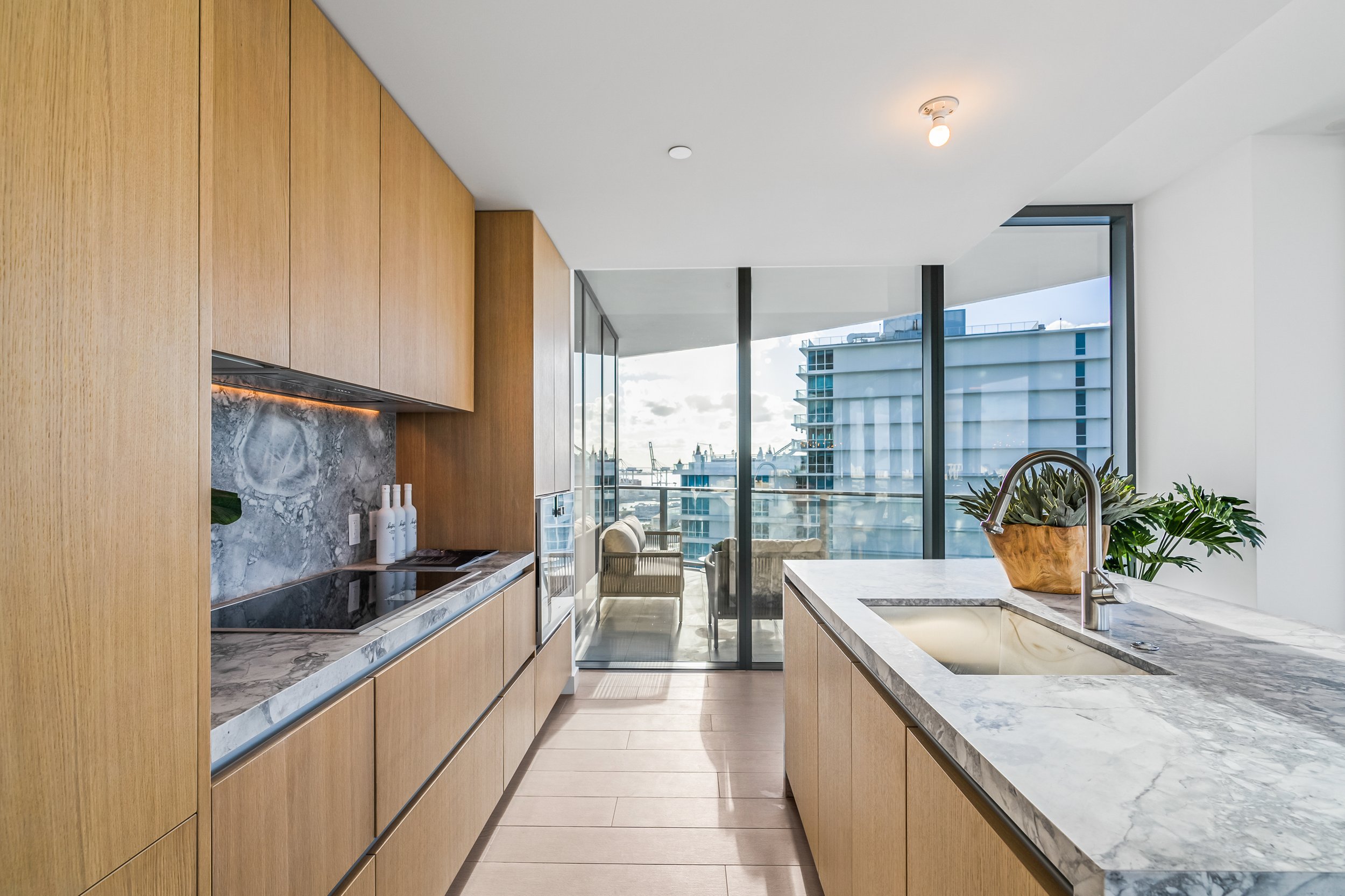Modern kitchen with wooden cabinets, marble countertops, and a view of a city through large windows.