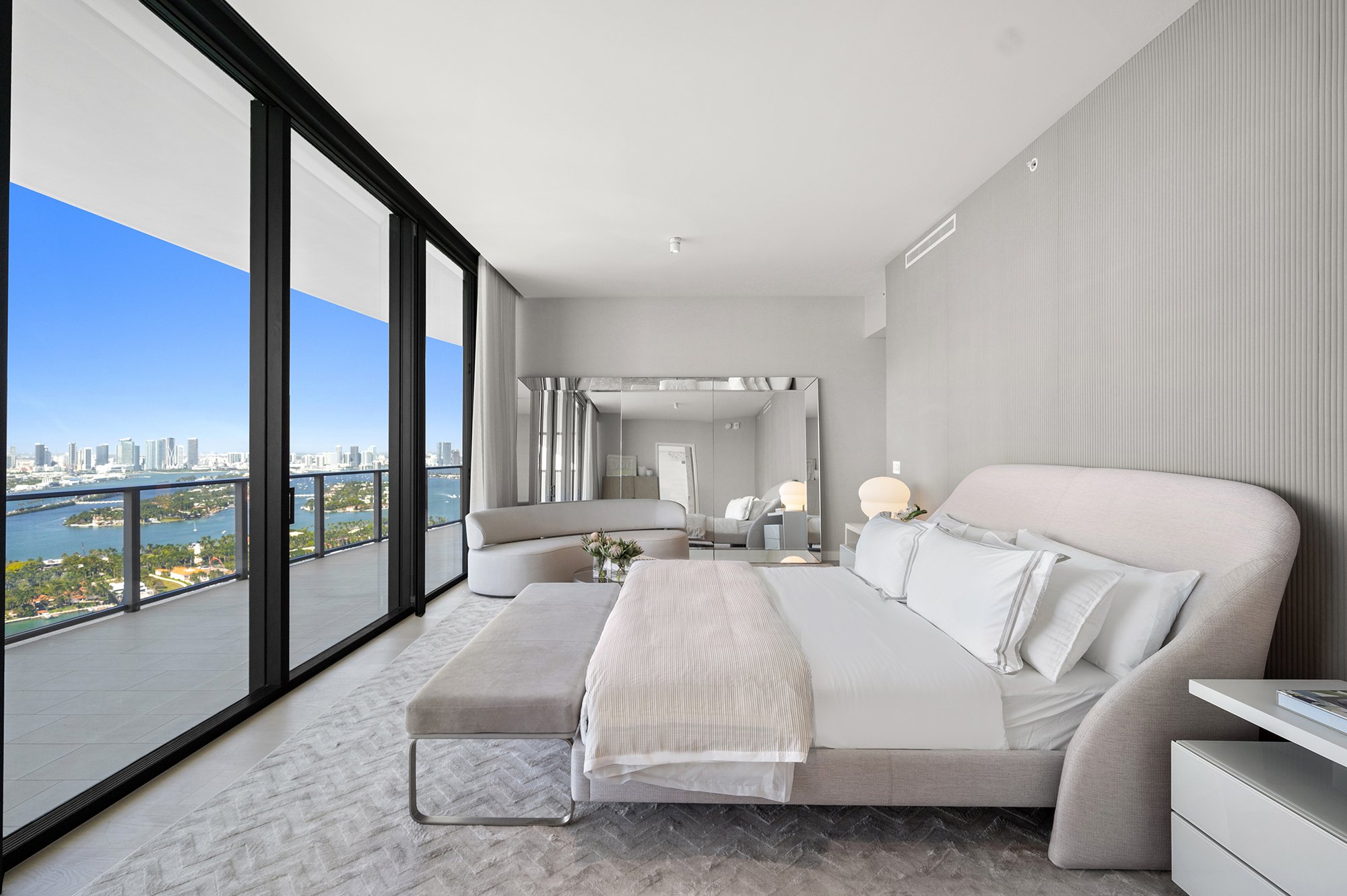 Modern bedroom with a large bed, gray upholstered headboard, and a bench at the foot. Floor-to-ceiling windows offer a view of a city skyline and waterfront. Neutral tones with minimalist decor, a mirrored wall, and a small seating area.