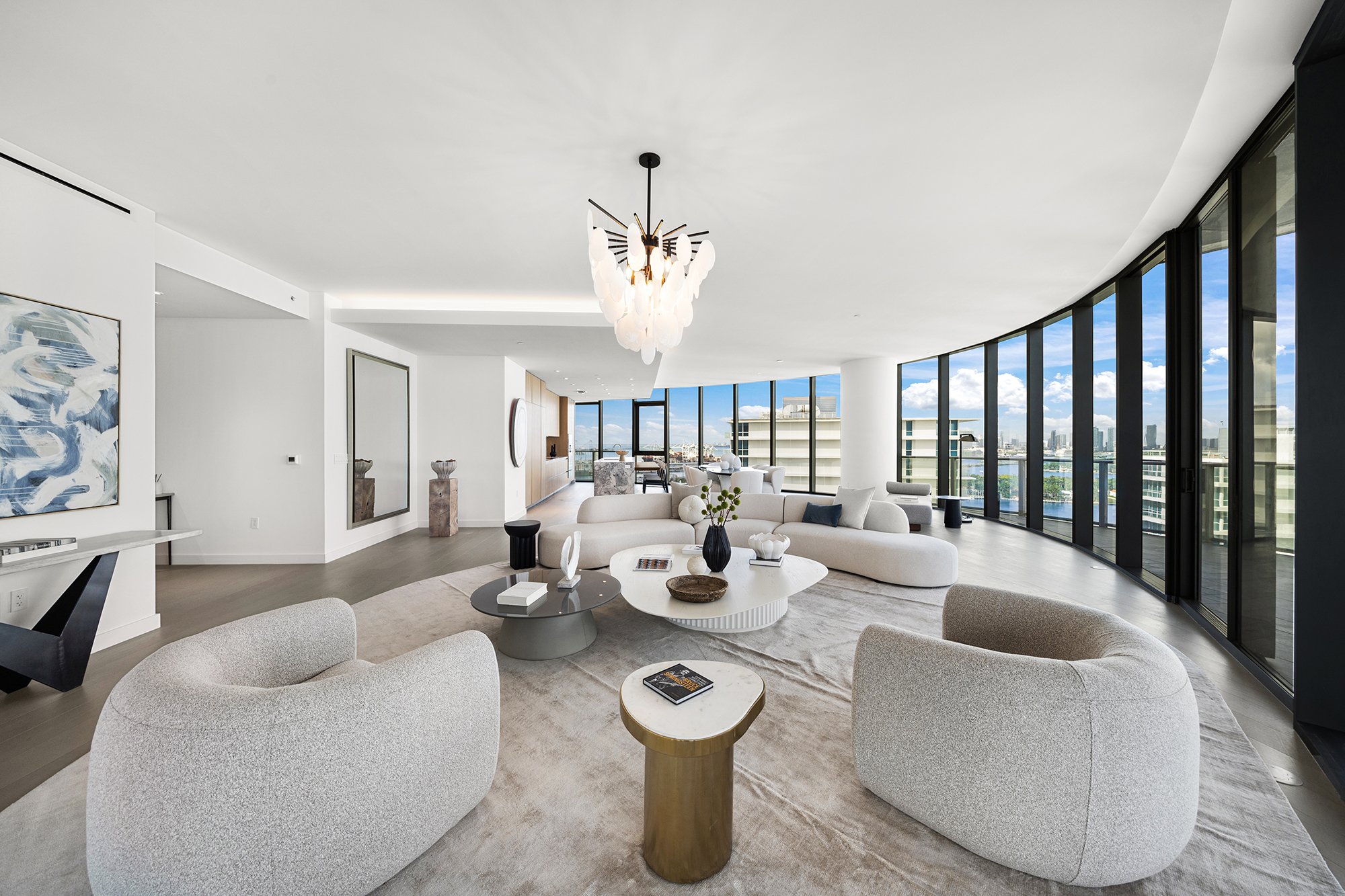 Modern open-concept living room with curved floor-to-ceiling windows, contemporary white sofas, an elegant chandelier, and a neutral color palette.