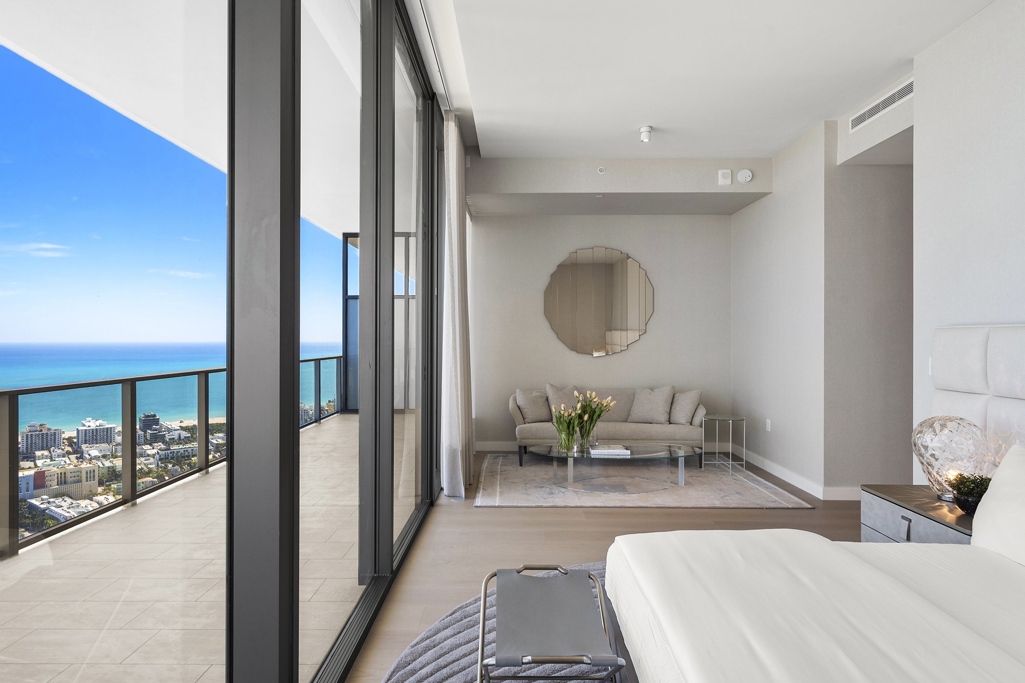 Modern bedroom with ocean view, large windows, white bed, gray couch, glass coffee table, decorative pillows, and plants, overlooking city skyline.