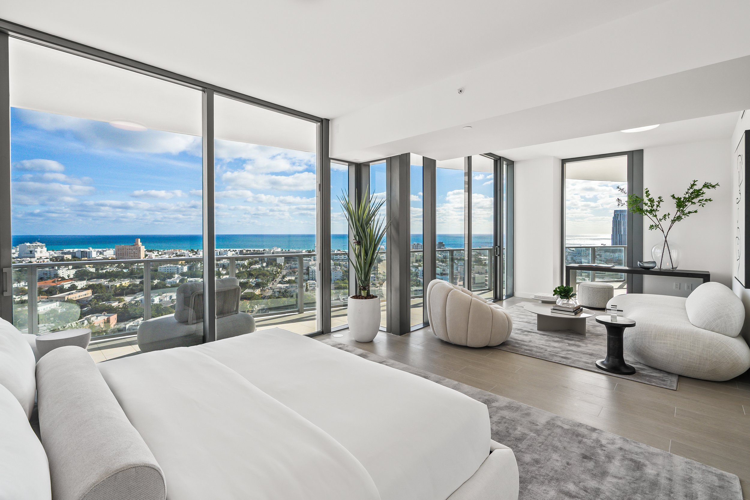 Luxury bedroom with modern decor, featuring a large bed, plush seating, floor-to-ceiling windows, and a stunning ocean view. The room has neutral tones, contemporary furniture, and indoor plants.