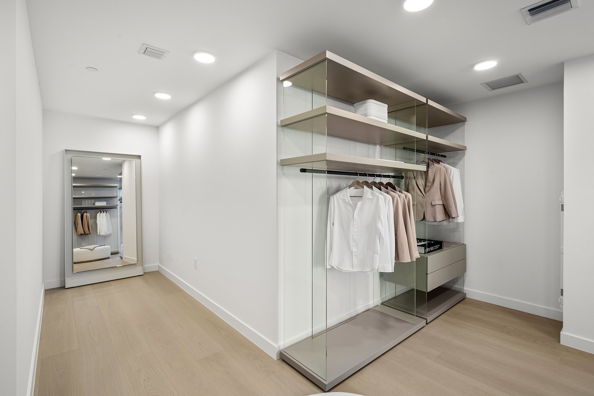 Modern walk-in closet with wooden floors, glass shelves, and clothes hanging neatly. A large mirror reflects the space, enhancing the room's brightness and spaciousness.