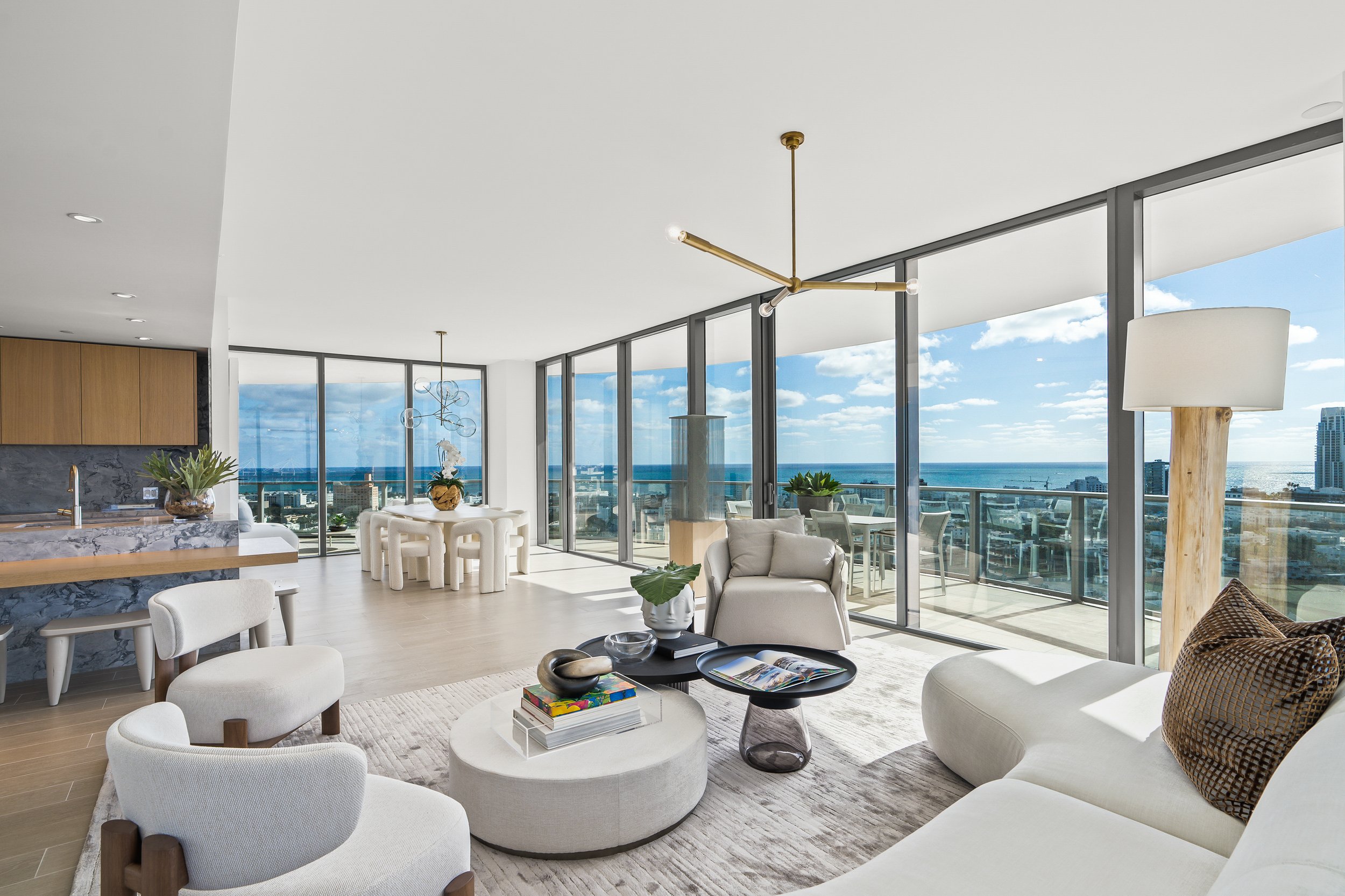Modern luxury living room with floor-to-ceiling windows, ocean view, contemporary furniture, and open layout. Features include a large white sectional sofa, round coffee table, neutral color palette, pendant lights, and adjacent dining area with a gl