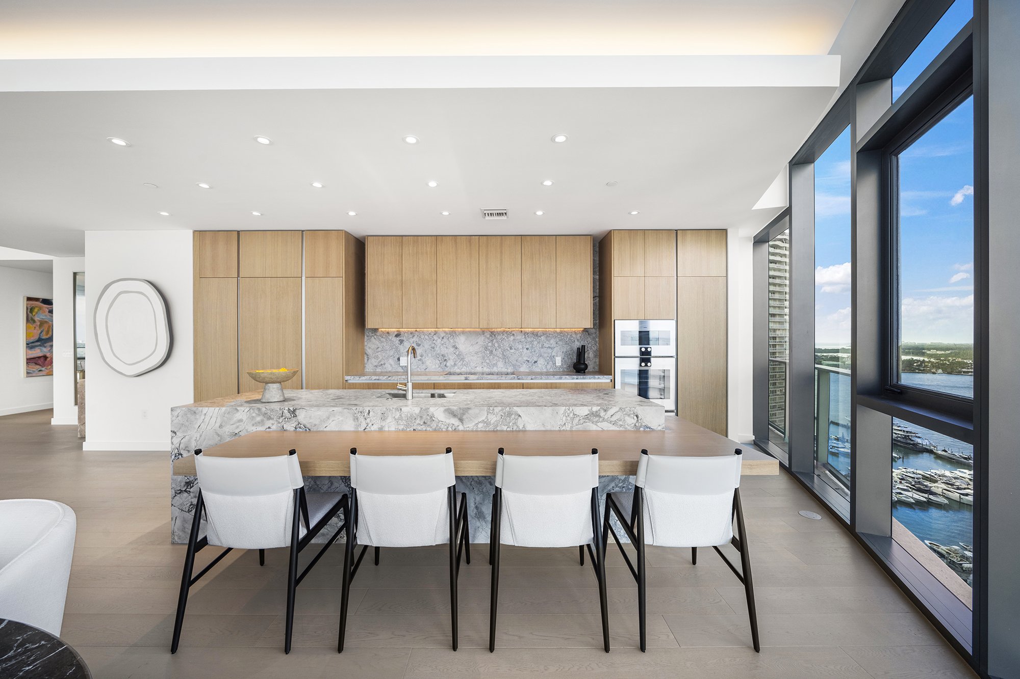 Modern kitchen with marble island, wooden cabinetry, and large windows overlooking a waterfront view.