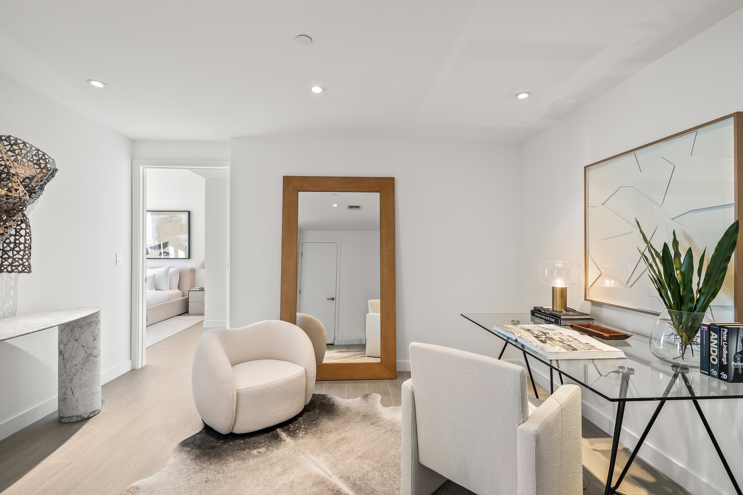 Modern home office with a glass desk, a chair, a large wooden-framed mirror, and abstract artwork. A marble console table is against the wall and a comfortable armchair is placed on a light-colored rug. There's also a plant and a decorative sculpture