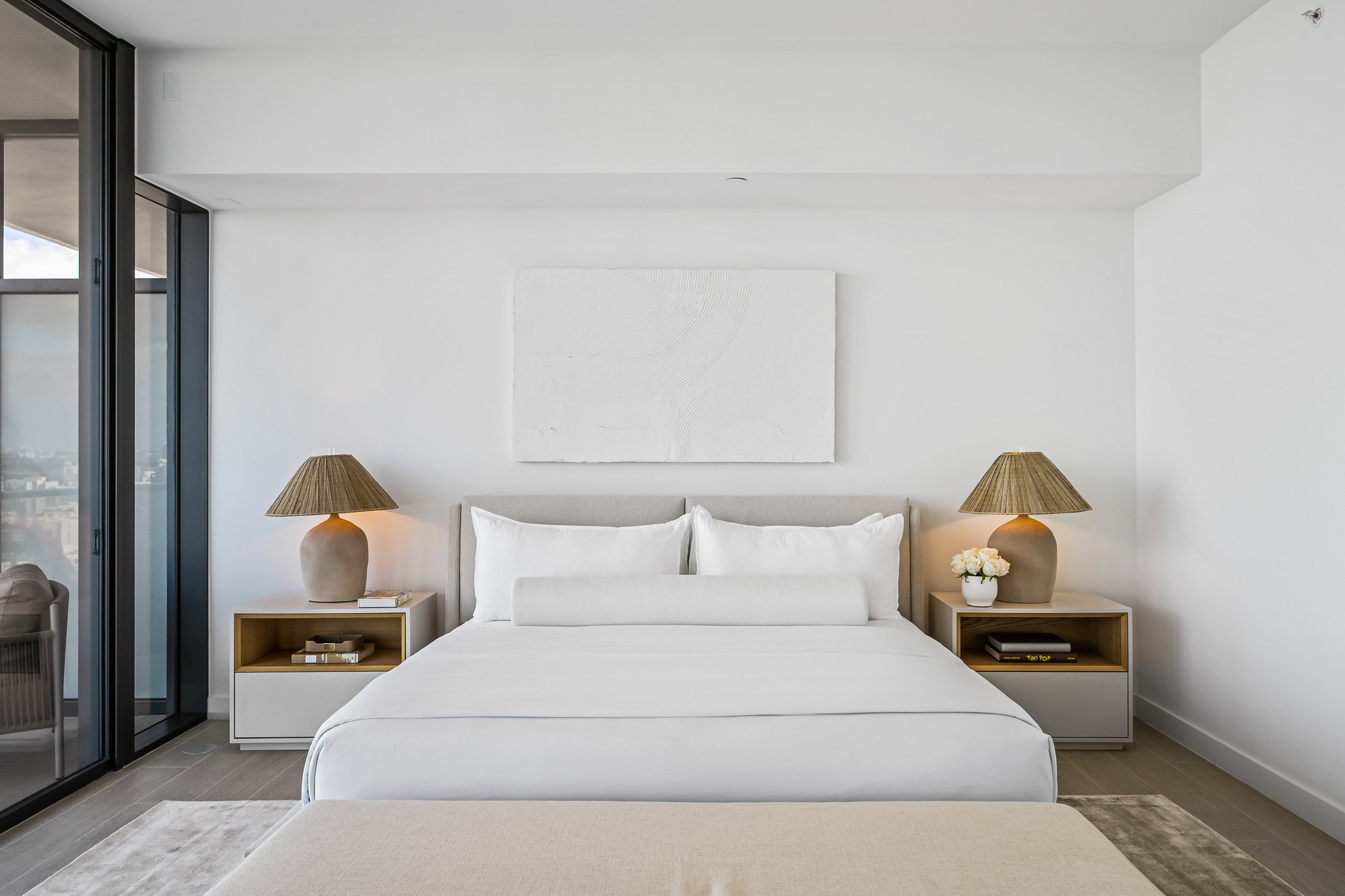 Modern bedroom with neutral decor, featuring a large bed with white linens, flanked by two nightstands each with a beige lamp, against a white wall with minimalist artwork.