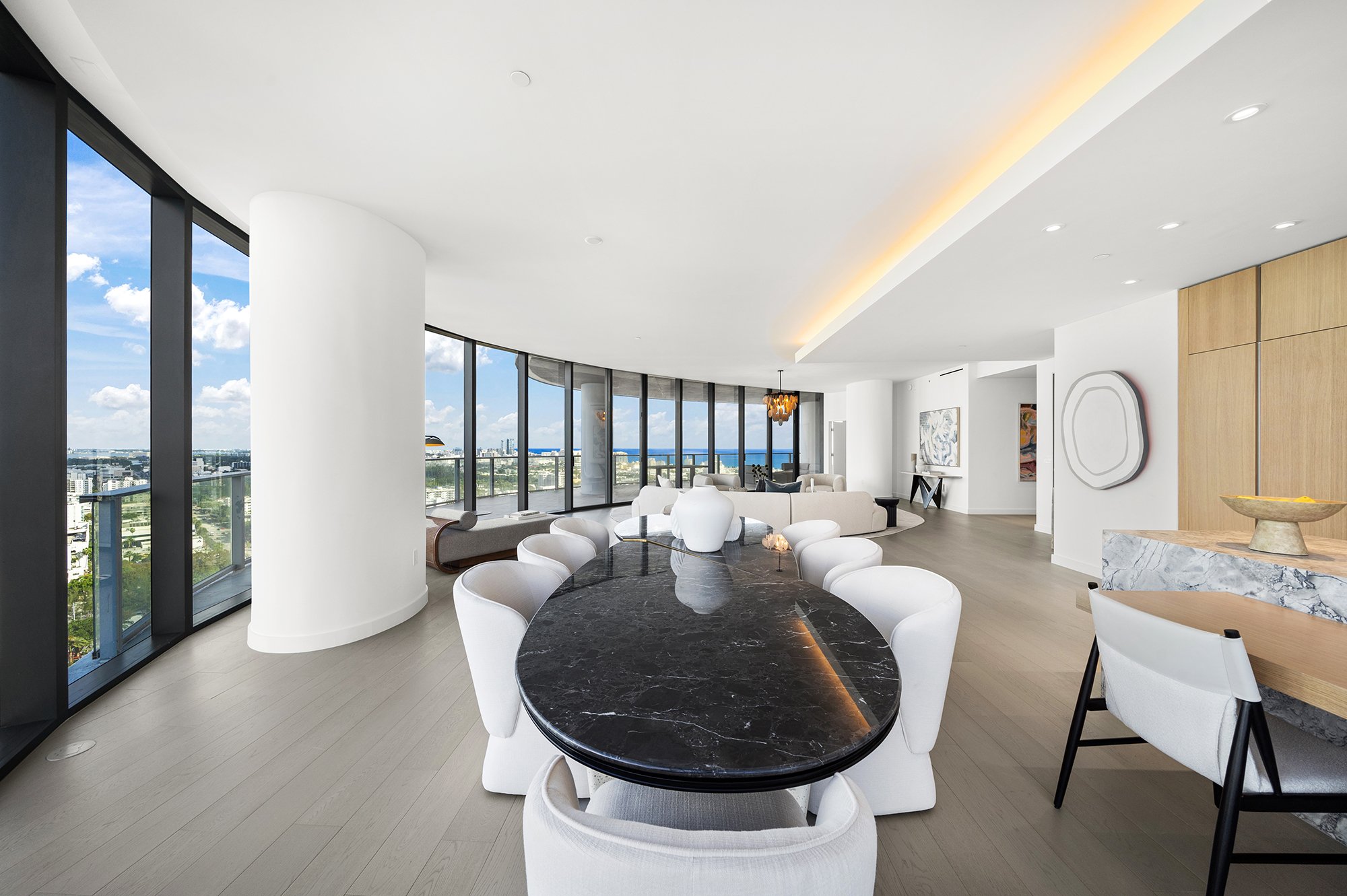 Spacious modern living area with large windows and city view, featuring round black marble dining table with white chairs, light wood flooring, and contemporary decor.