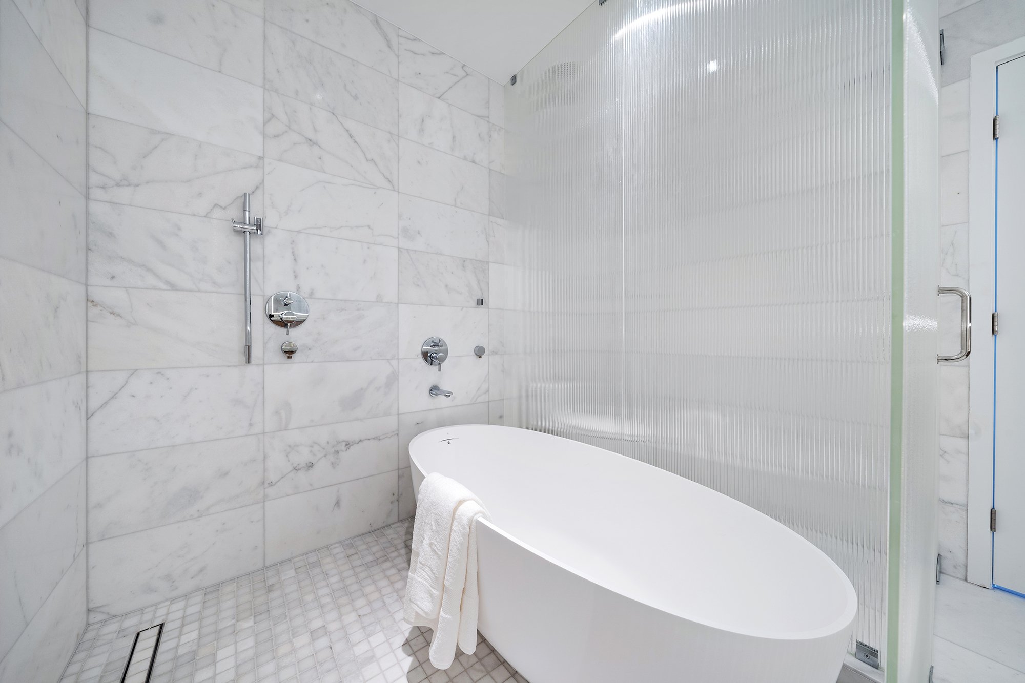 Modern bathroom with a white freestanding bathtub, marble tile walls, glass shower enclosure, and chrome fixtures.