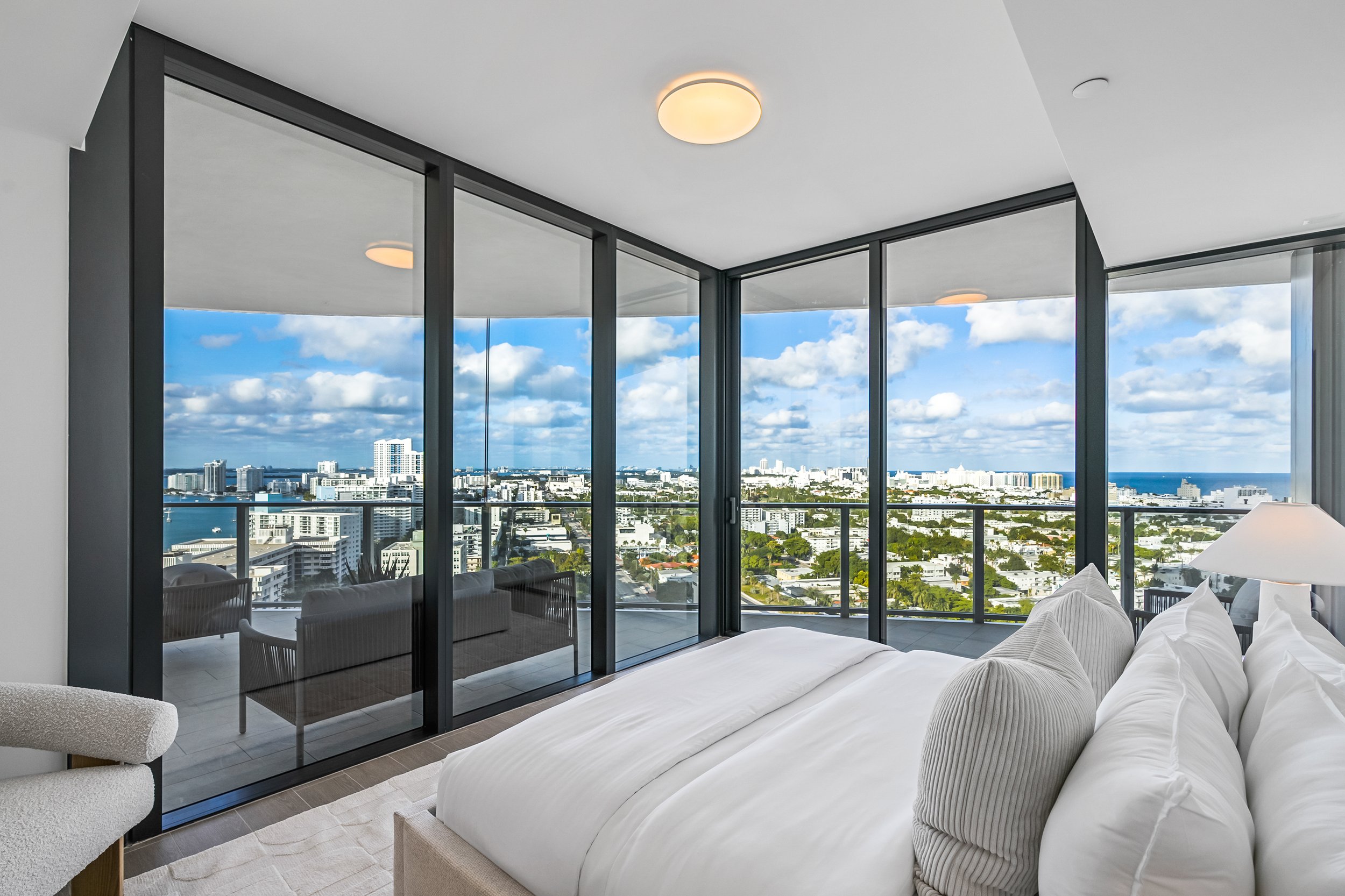 Luxurious bedroom with floor-to-ceiling glass windows offering a panoramic city skyline and ocean view, featuring a bed with white bedding and a soft chair.