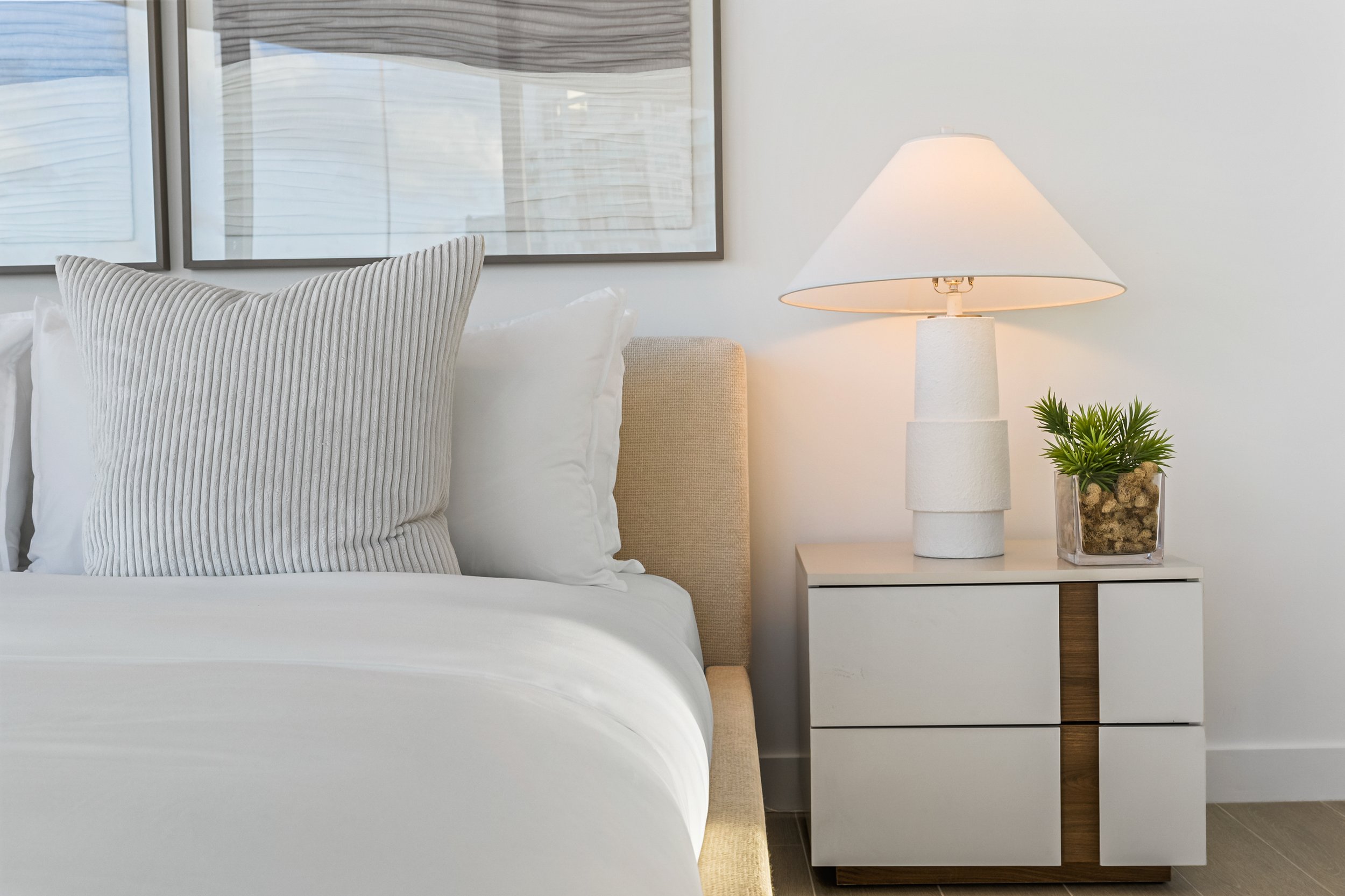Modern bedroom with a bedside table, lamp, plant, and art on the wall. A bed with white sheets and a textured pillow is visible.