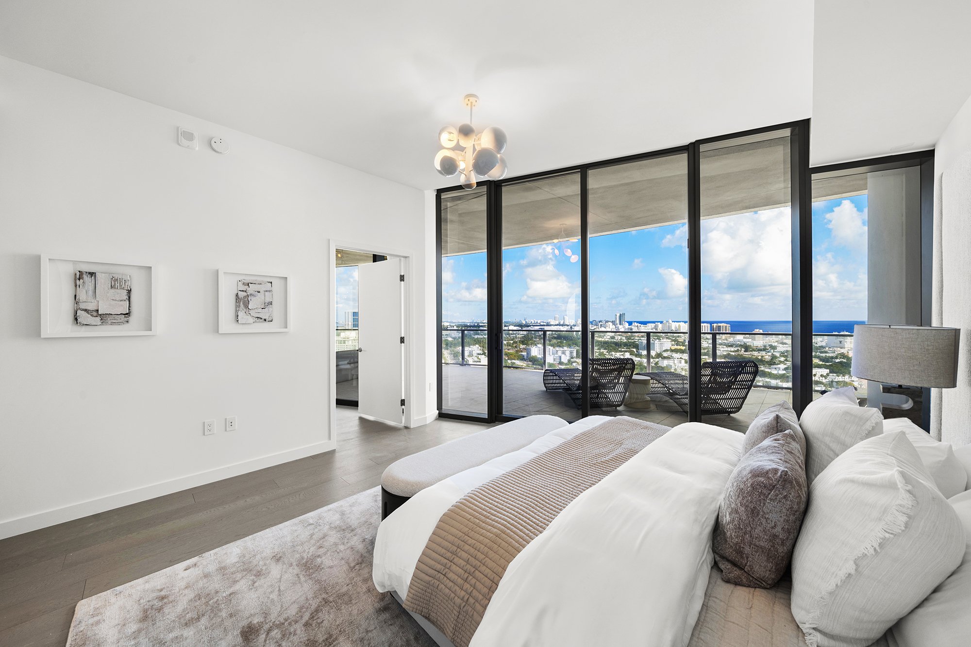 Modern bedroom interior with a large bed, pillows, floor-to-ceiling glass windows leading to a balcony, and city skyline view.