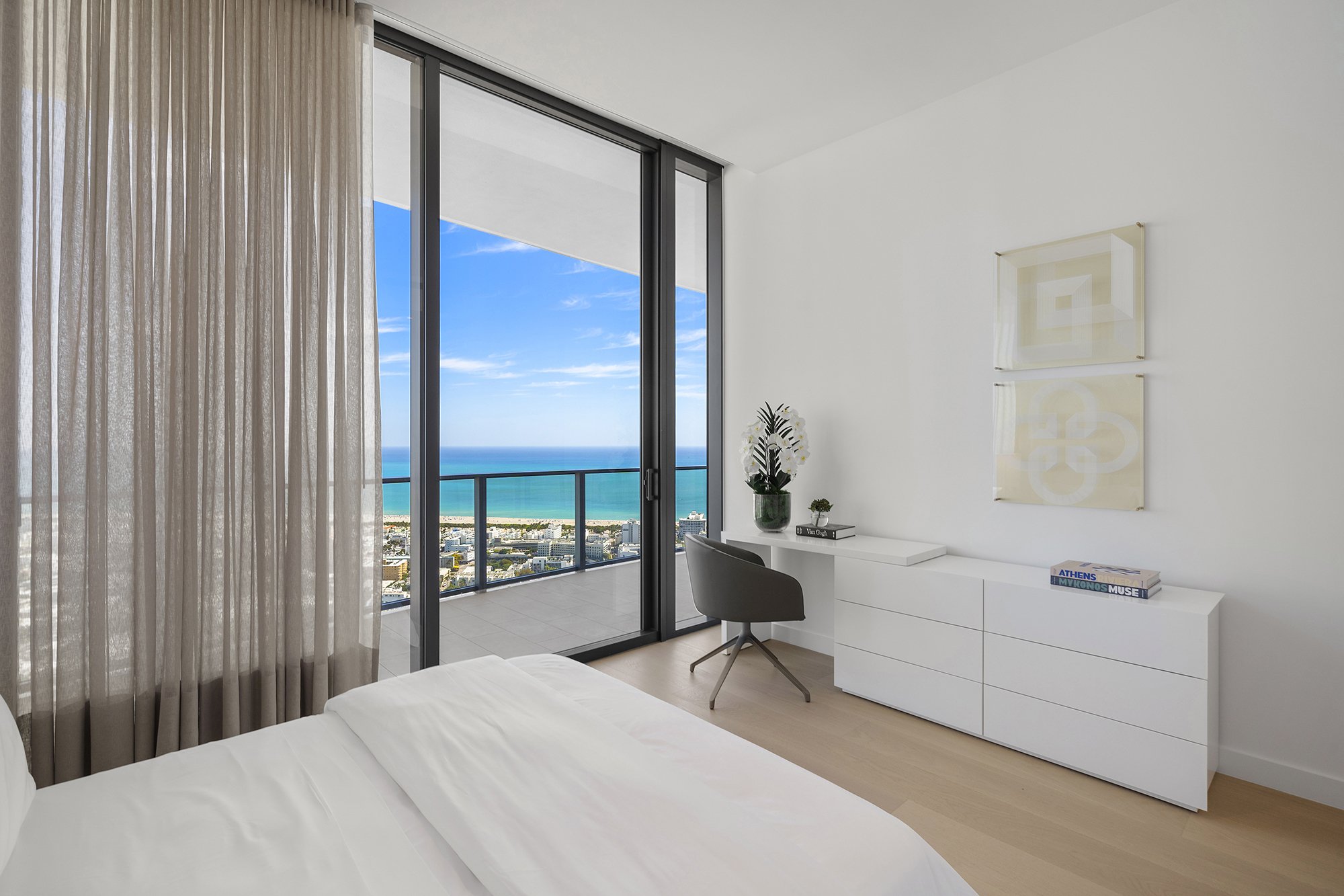 Modern bedroom with a white bed, glass sliding doors, ocean view, desk with chair, and minimal decor.
