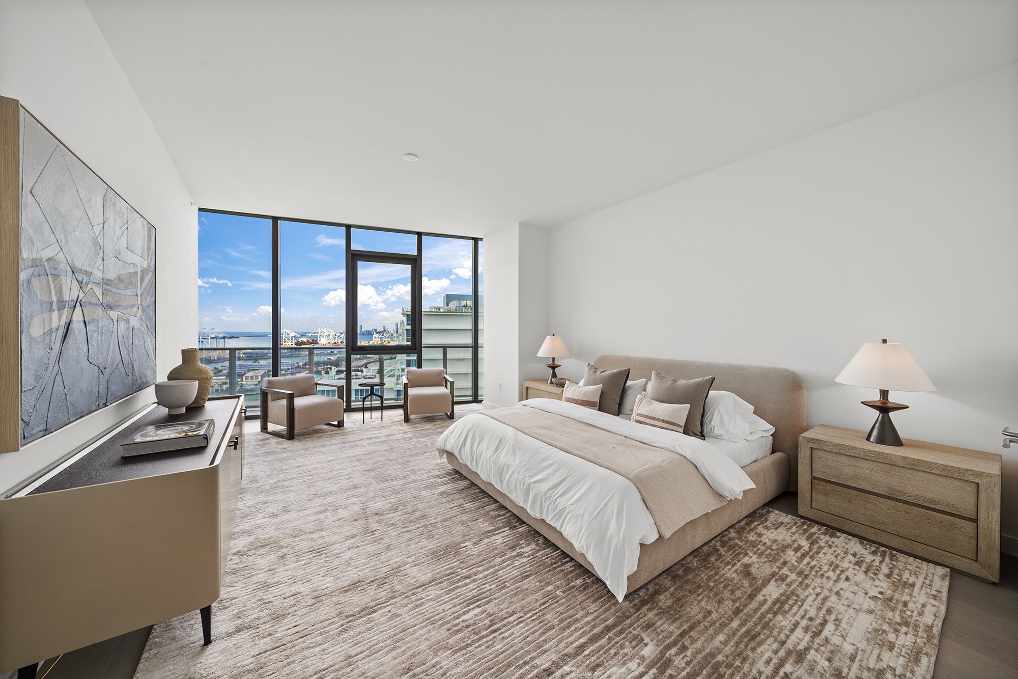 Modern bedroom with large windows, king-size bed, beige decor, and city view.