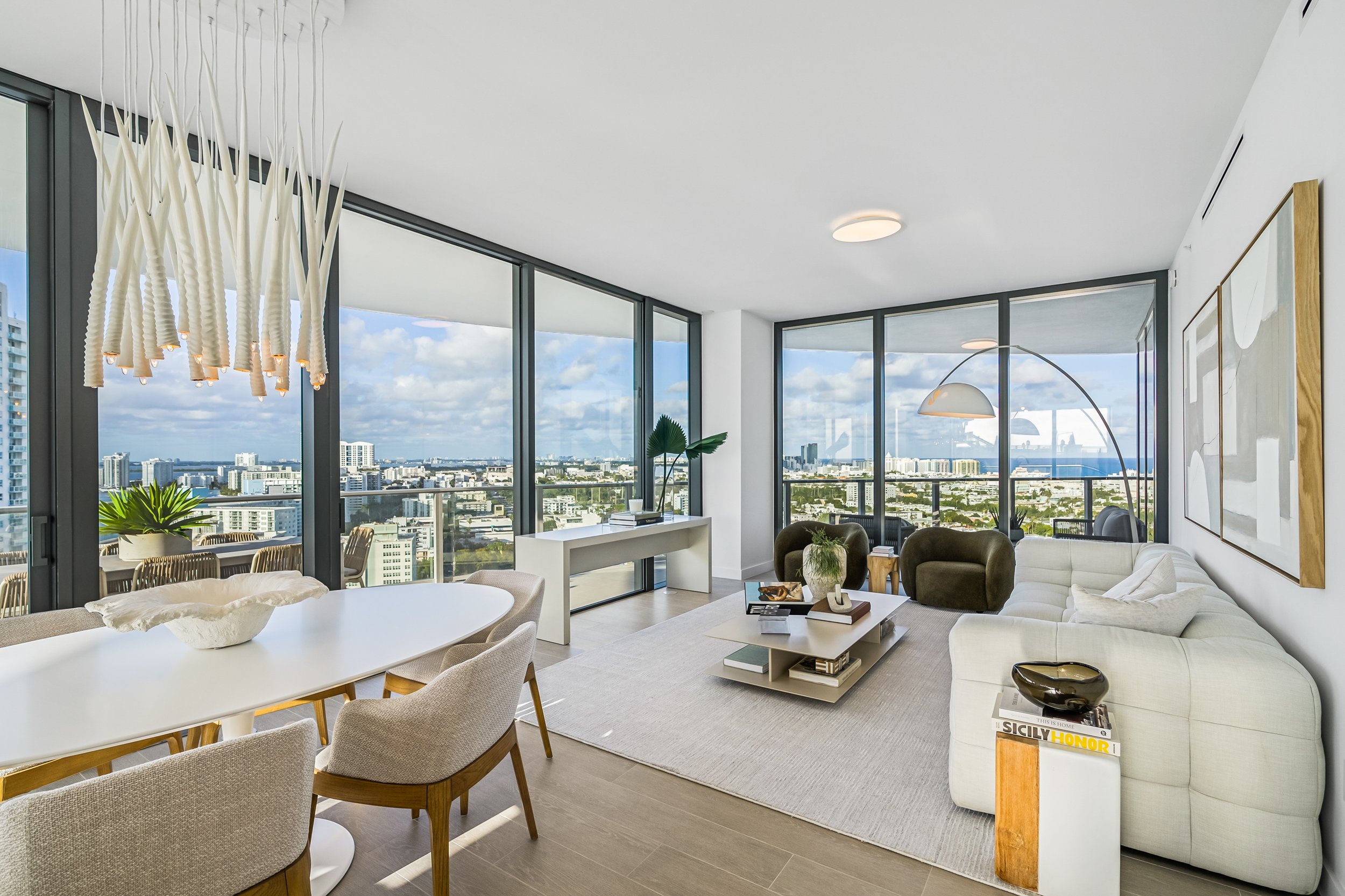 Modern living and dining area with panoramic city views, featuring a white dining table, plush seating, and contemporary decor. Floor-to-ceiling windows allow natural light to flood the space. A unique chandelier hangs over the dining area.