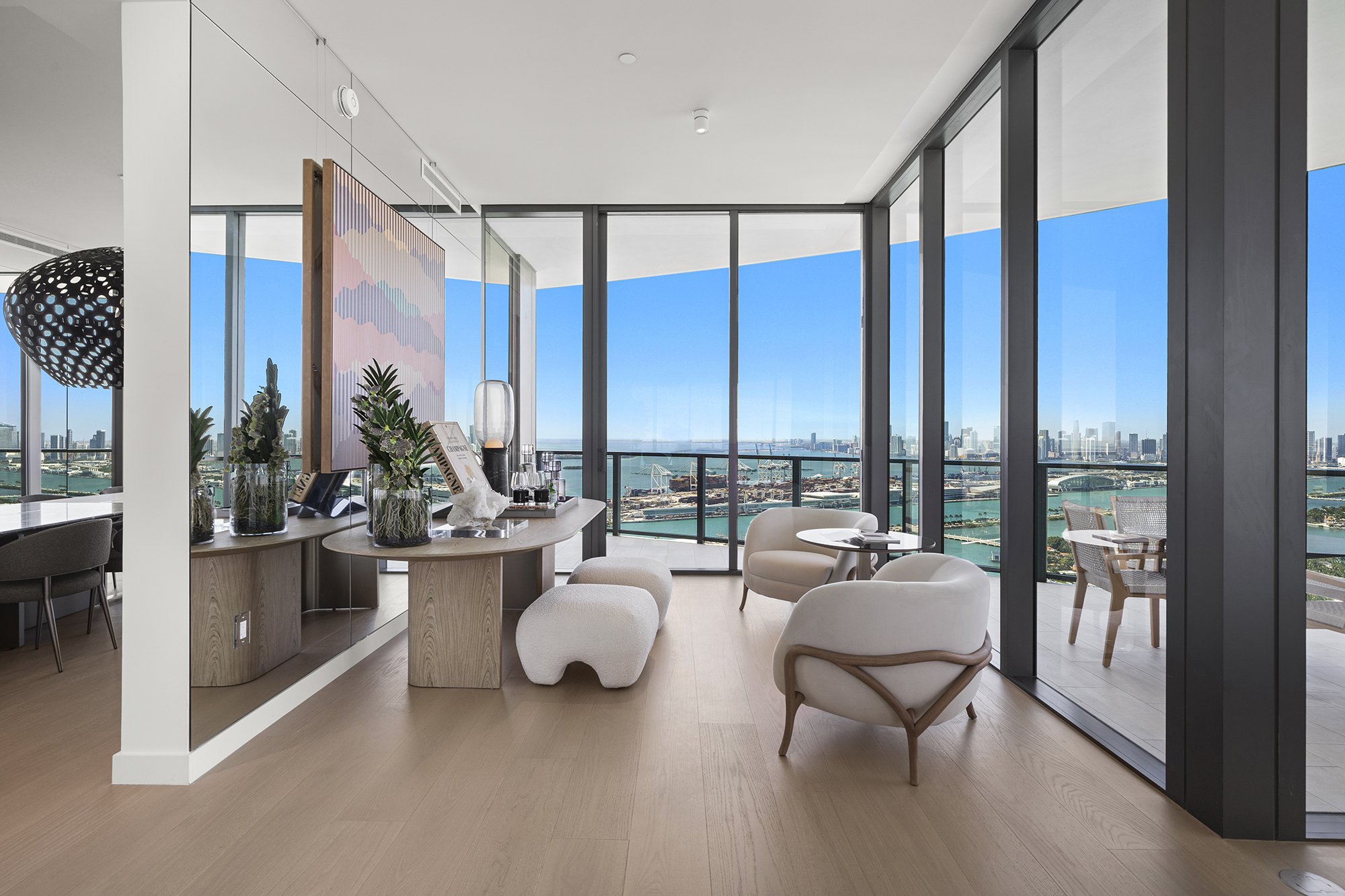 Modern living room with large windows and cityscape view, featuring contemporary furniture and decor, including armchairs, a small round table, potted plants, and a colorful abstract artwork.