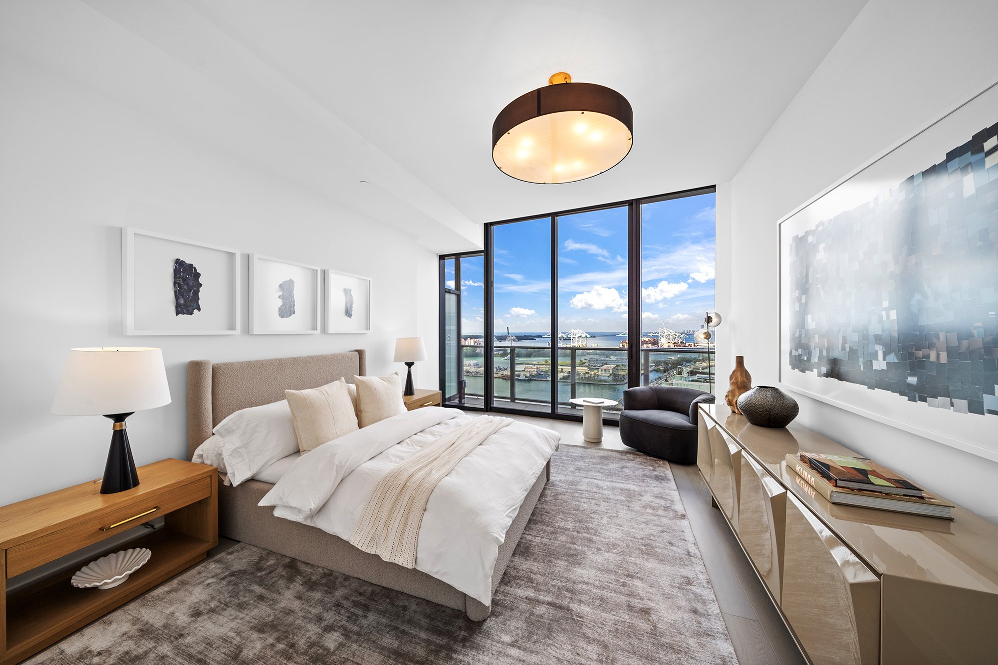 Modern bedroom with large window view of city harbor, featuring a bed with beige bedding, nightstands with lamps, abstract wall art, and a light fixture. Room includes a small seating area and decorative elements on a sideboard.