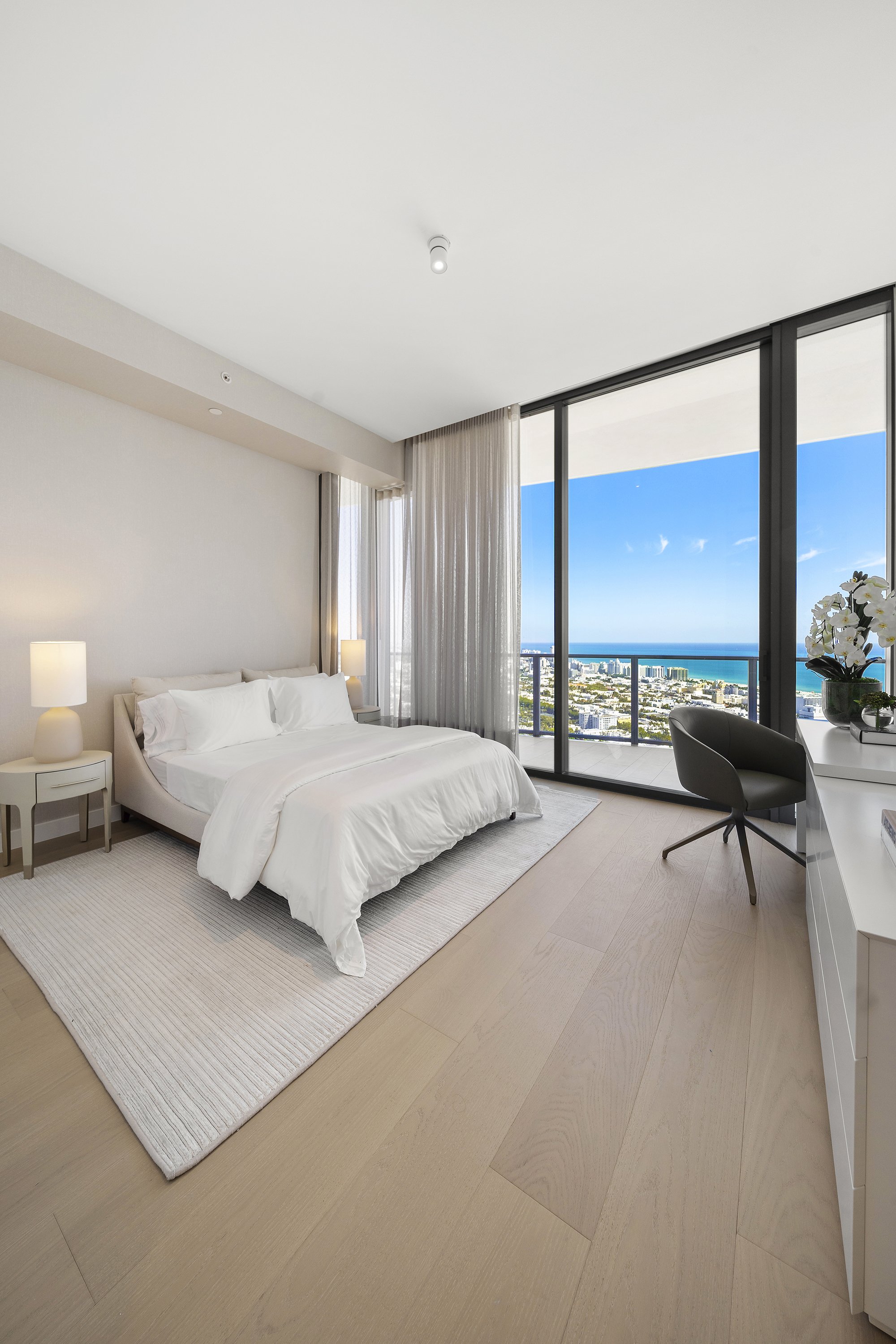 Modern bedroom with a large bed, white bedding, floor-to-ceiling windows, wooden floor, desk with a chair, and ocean view.