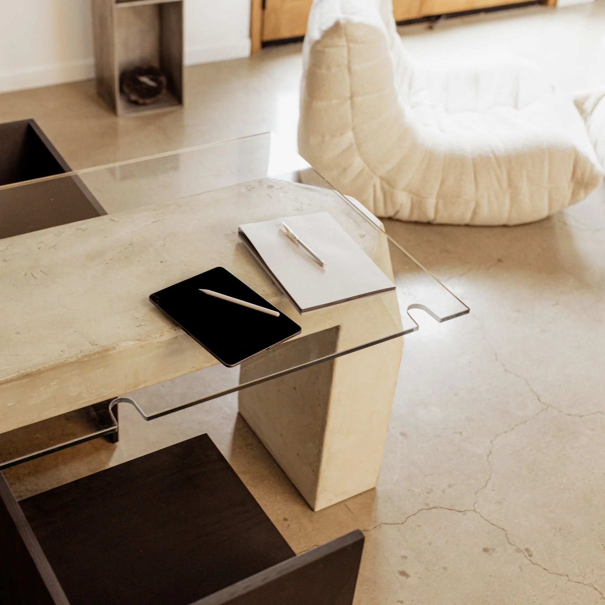 A modern, minimalist desk with a clear acrylic ledge, a black tablet with a white stylus, a notepad, and a pen. In the background, there is a cream-colored, plush armchair and wooden shelves.
