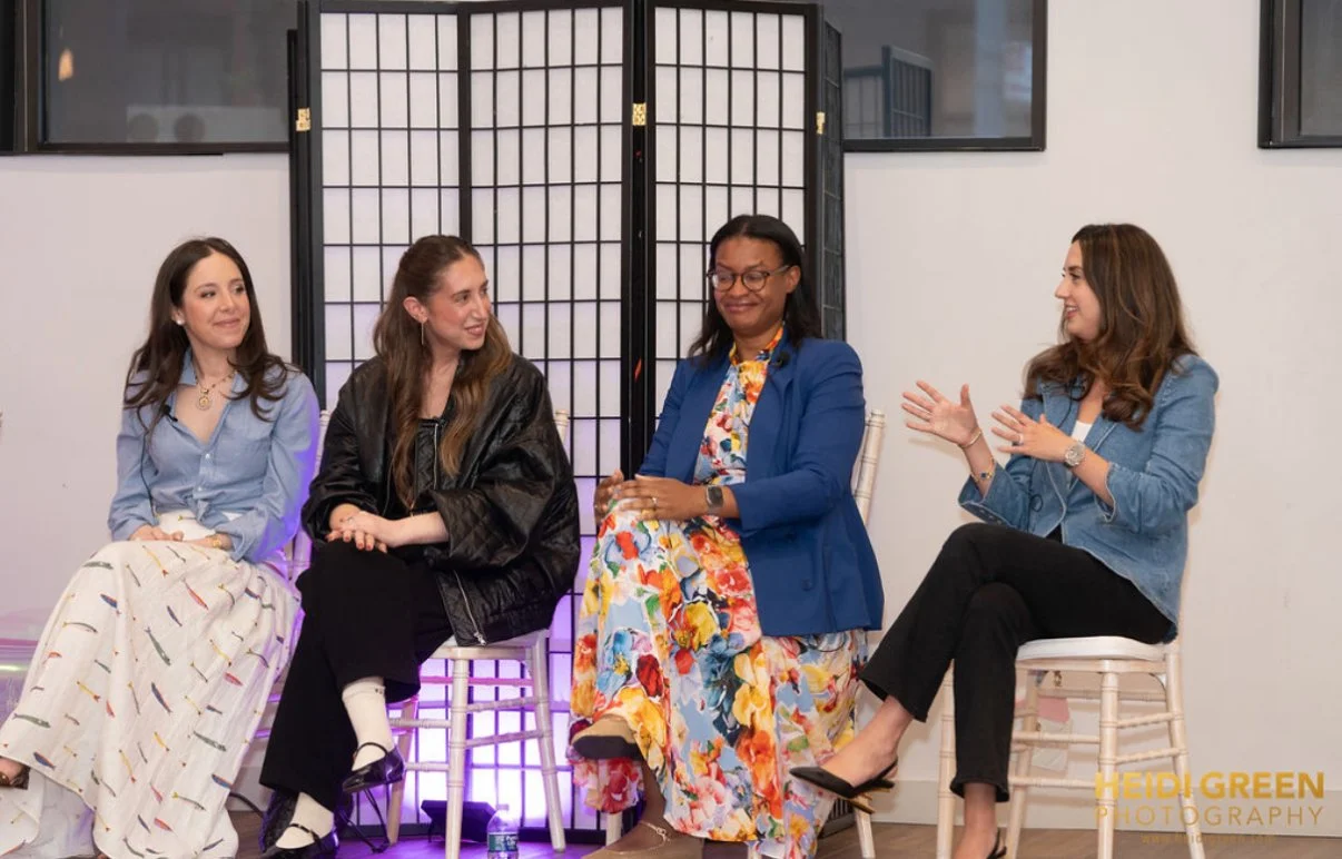 Four women sitting on chairs and engaging in conversation, with a folding room divider behind them, in an indoor setting.