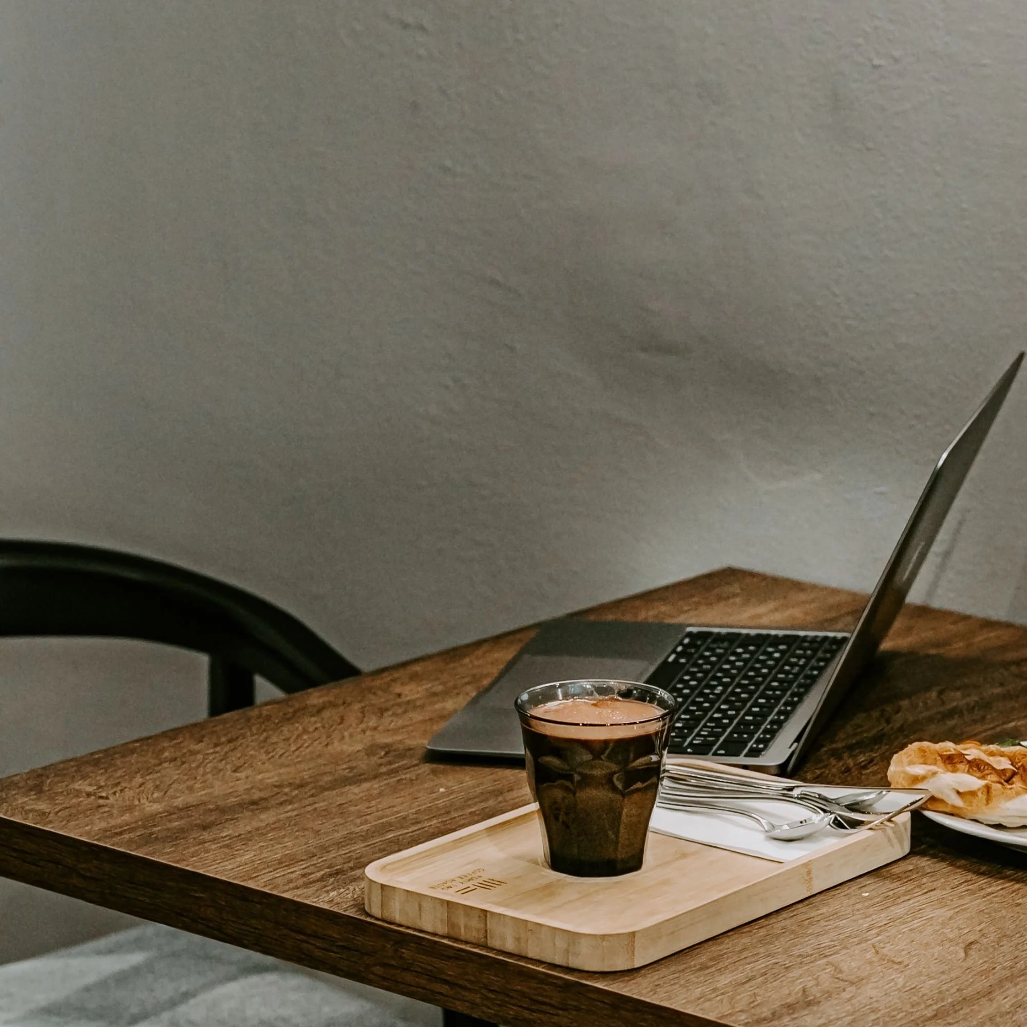 A laptop, a wooden tray with a glass of dark coffee, and a plate with a pastry on a wooden table.
