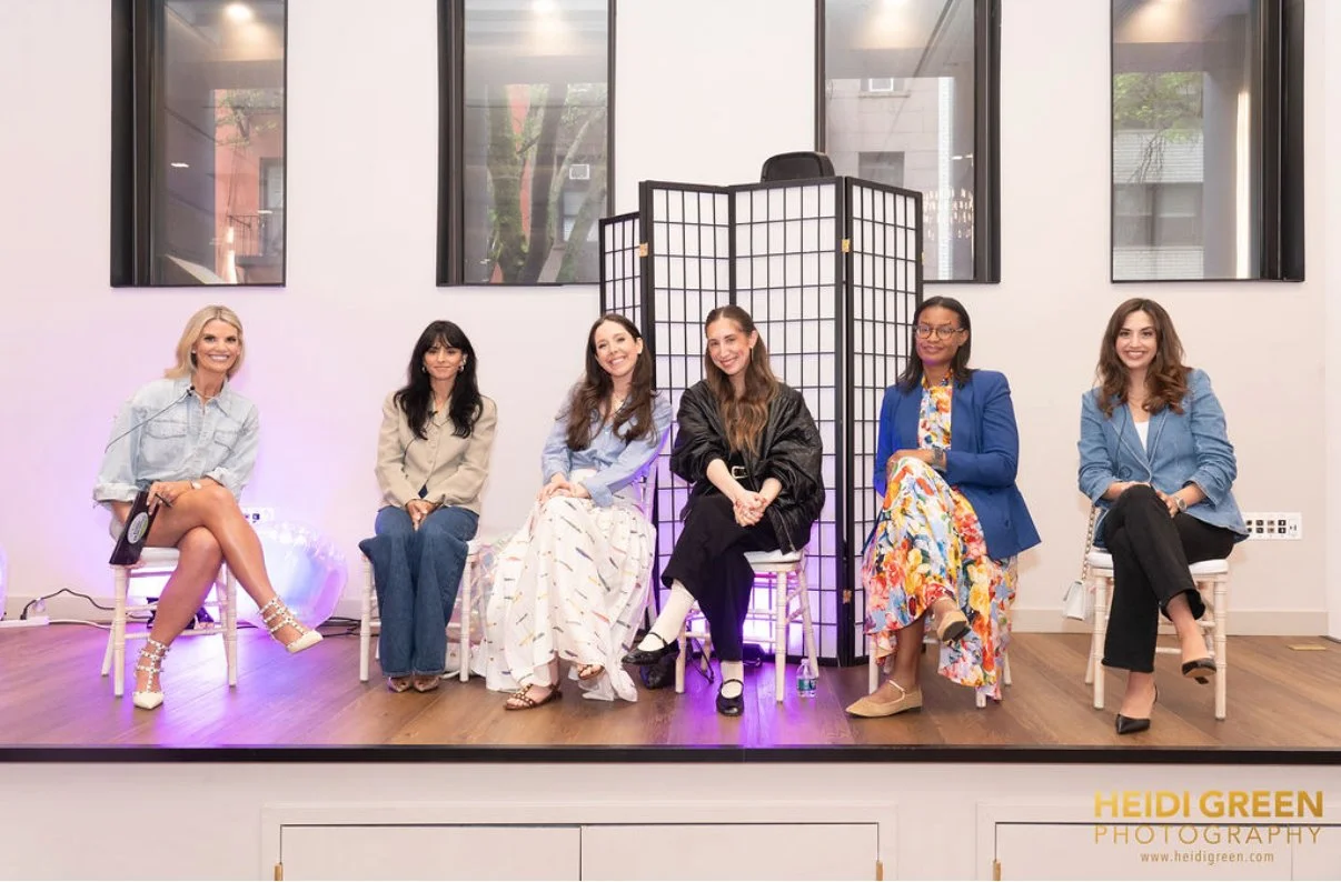 Six women sitting on chairs in a room with large windows and a folding screen behind them, participating in a panel or discussion event.