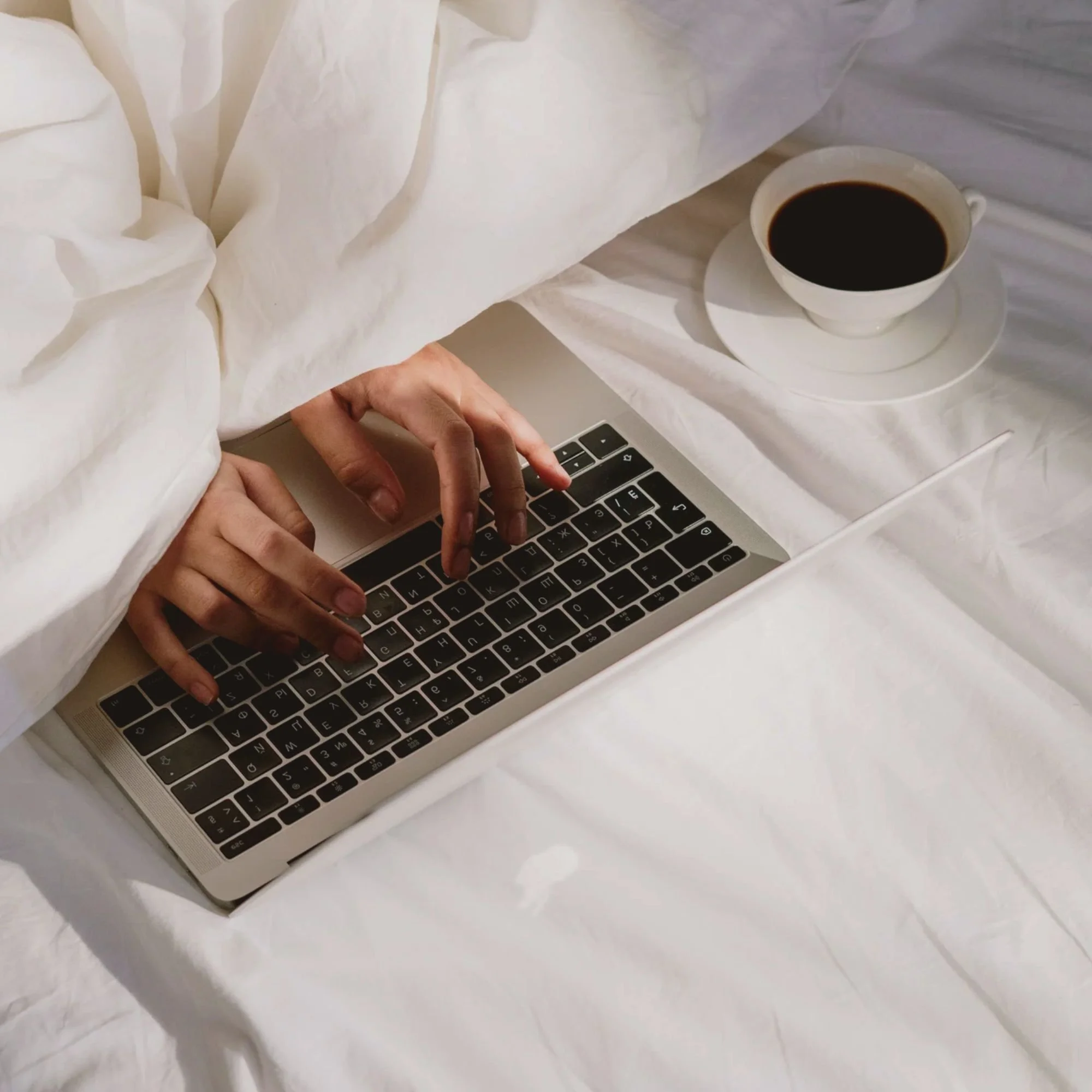 Person typing on a laptop under a white blanket, with a cup of coffee on a saucer nearby on a white bed.