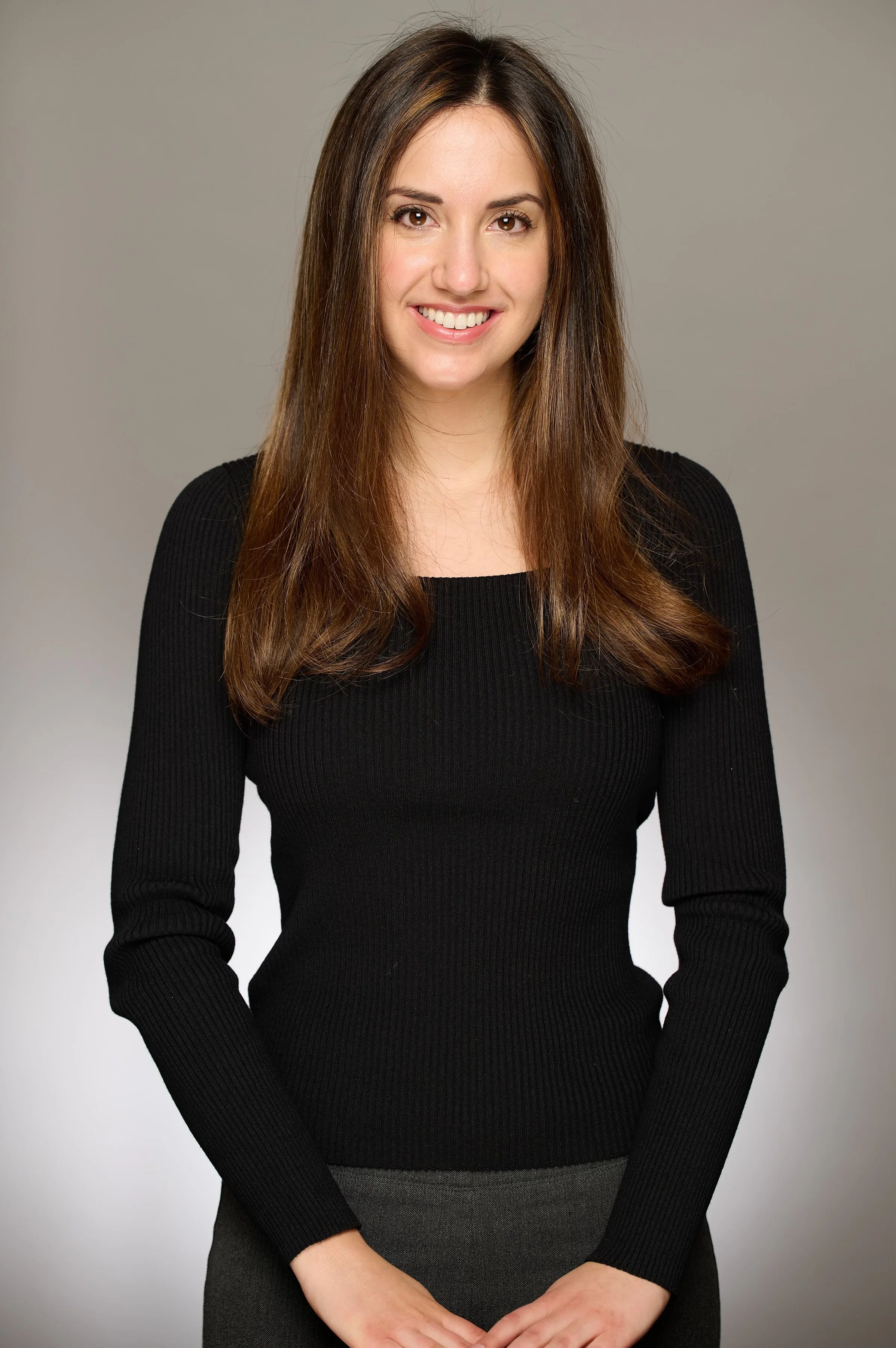 A young woman with long brown hair wearing a black ribbed long-sleeve top, smiling against a neutral gray background.