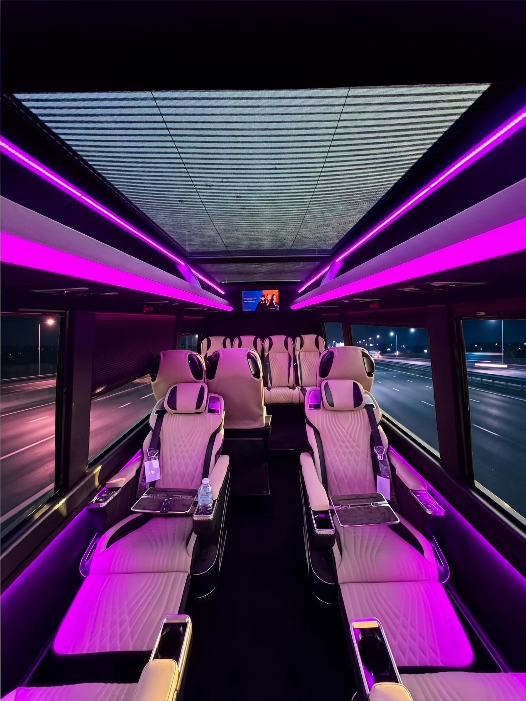 Interior of a modern bus or coach at night with pink and purple LED lighting, beige seats, large windows showing a highway, and a screen at the front displaying an advertisement.