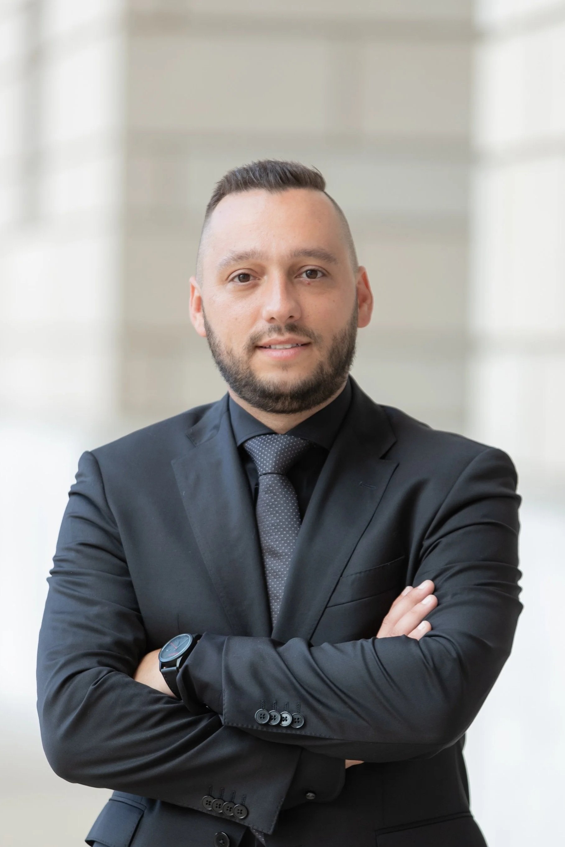 A man in a black suit and tie standing with arms crossed in a modern building with large windows.