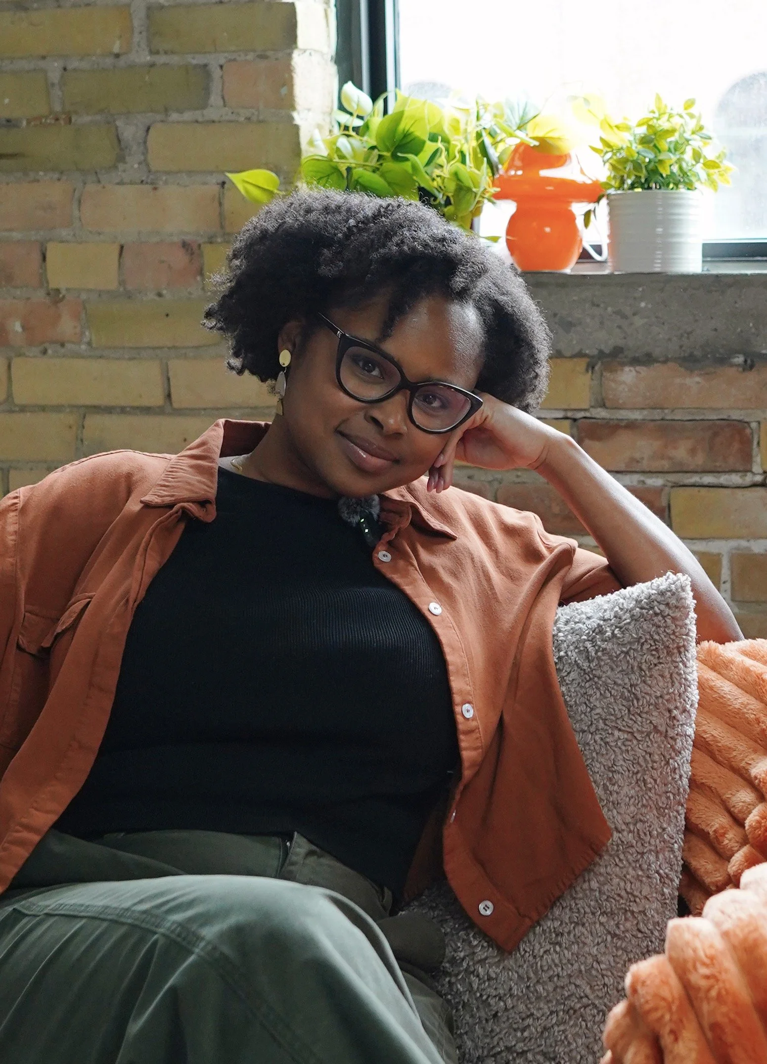 A woman with glasses, dark curly hair, and a light brown jacket sitting on a cozy gray and beige blanket, resting her head on her hand, in an interior space with a brick wall and potted plants on a window ledge.