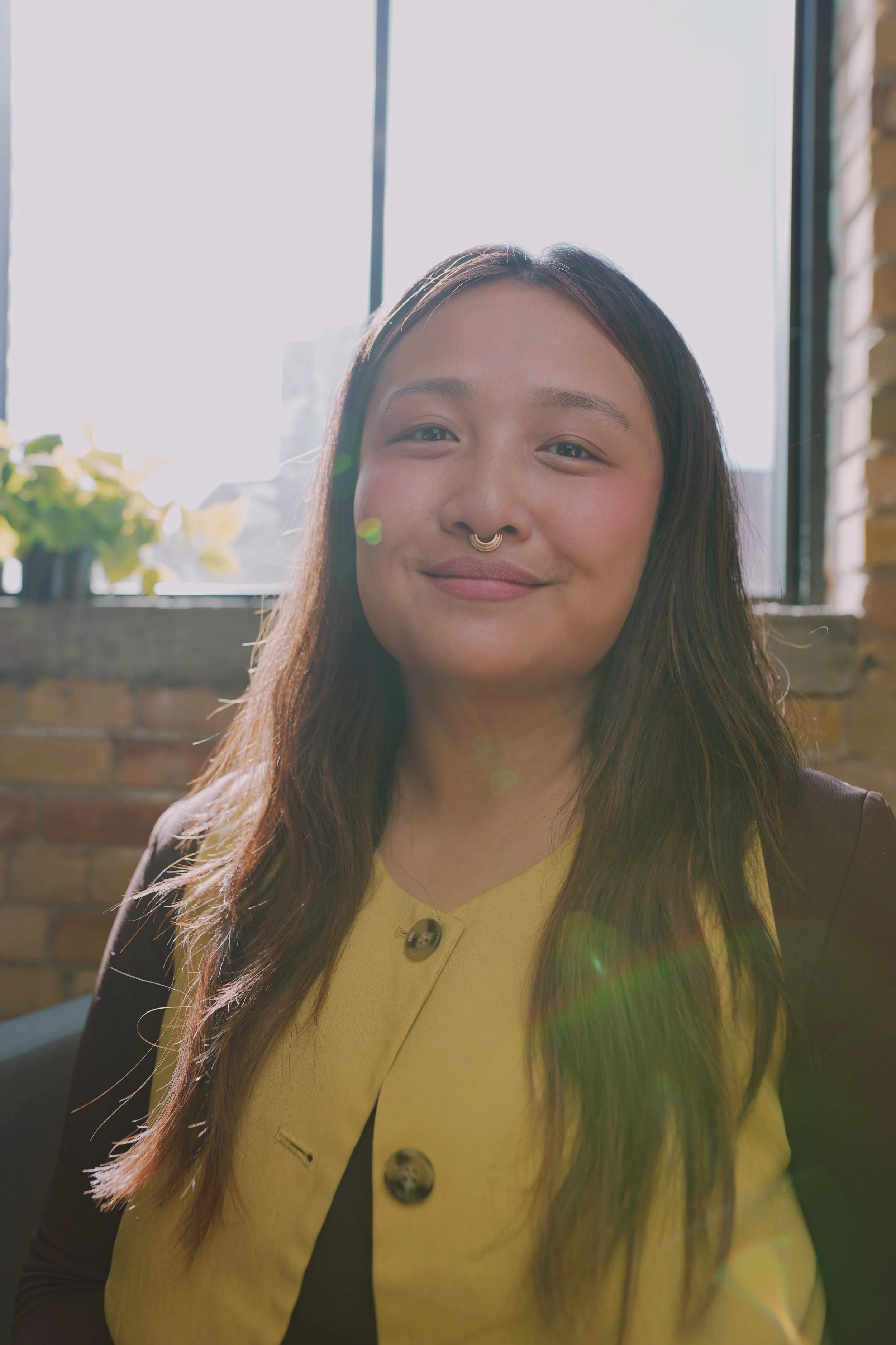 Young woman with long hair, smiling, wearing a yellow top and a dark jacket, standing indoors near a window with sunlight and a brick wall background.