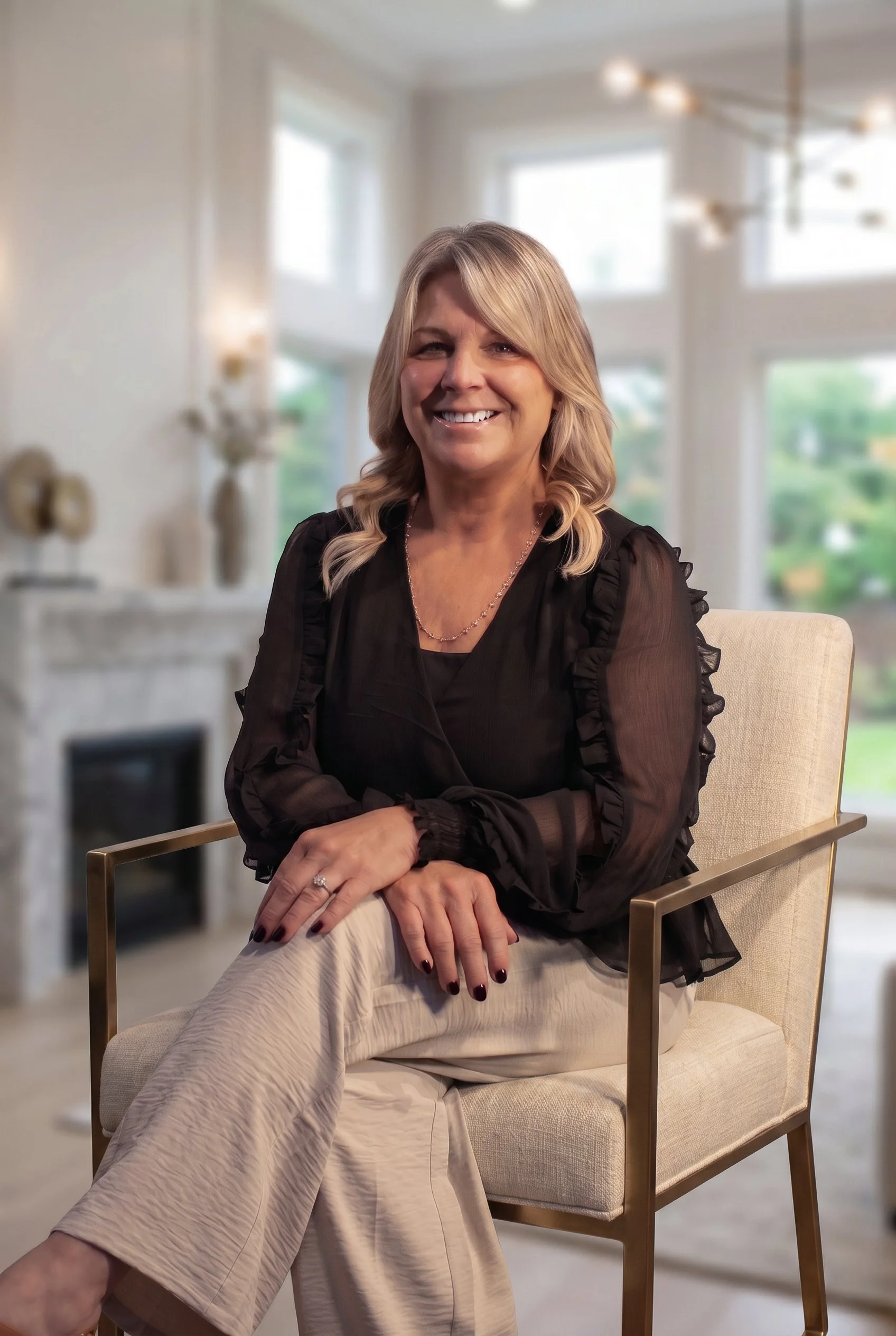 A smiling woman with blonde hair sitting in a modern, light-filled living room with large windows in the background, wearing a black blouse and beige pants.
