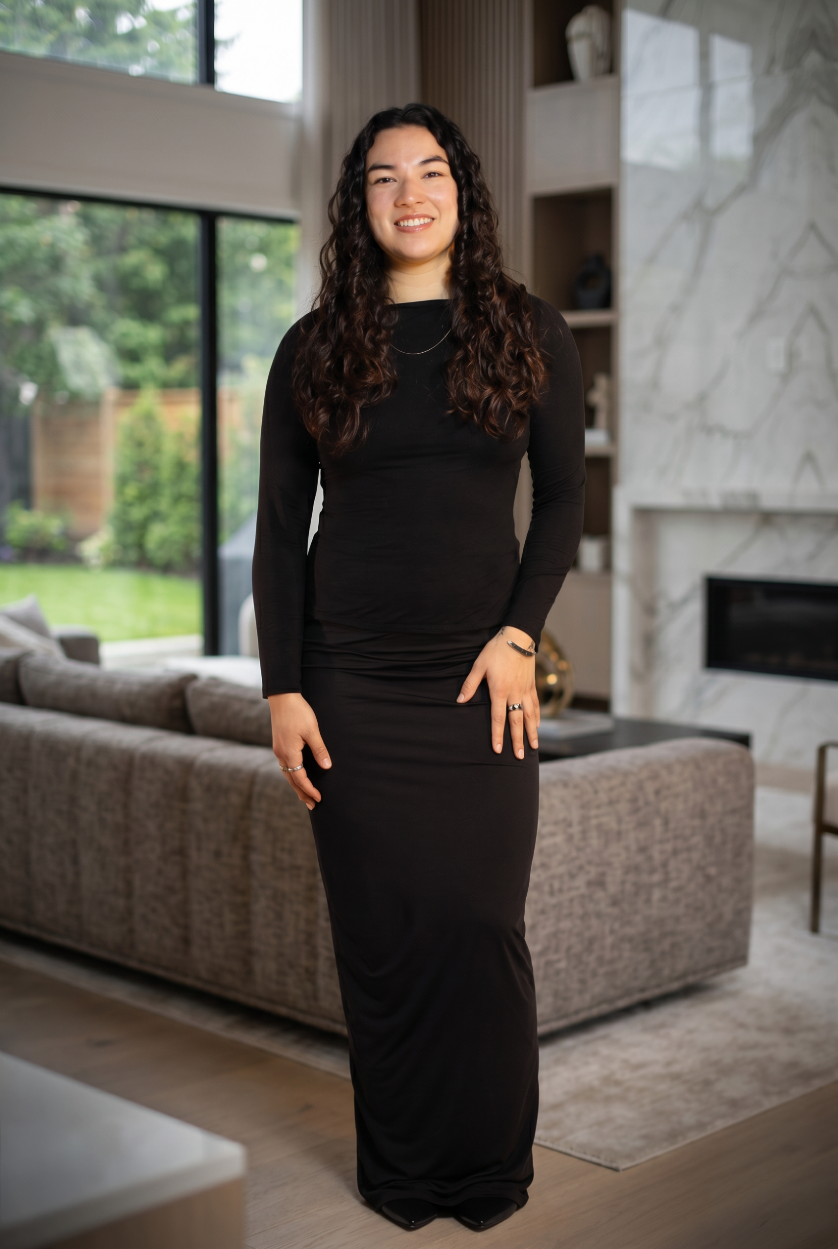 A woman with long curly hair, wearing a black dress, standing in a modern living room with large windows, a beige sofa, and a marble fireplace.
