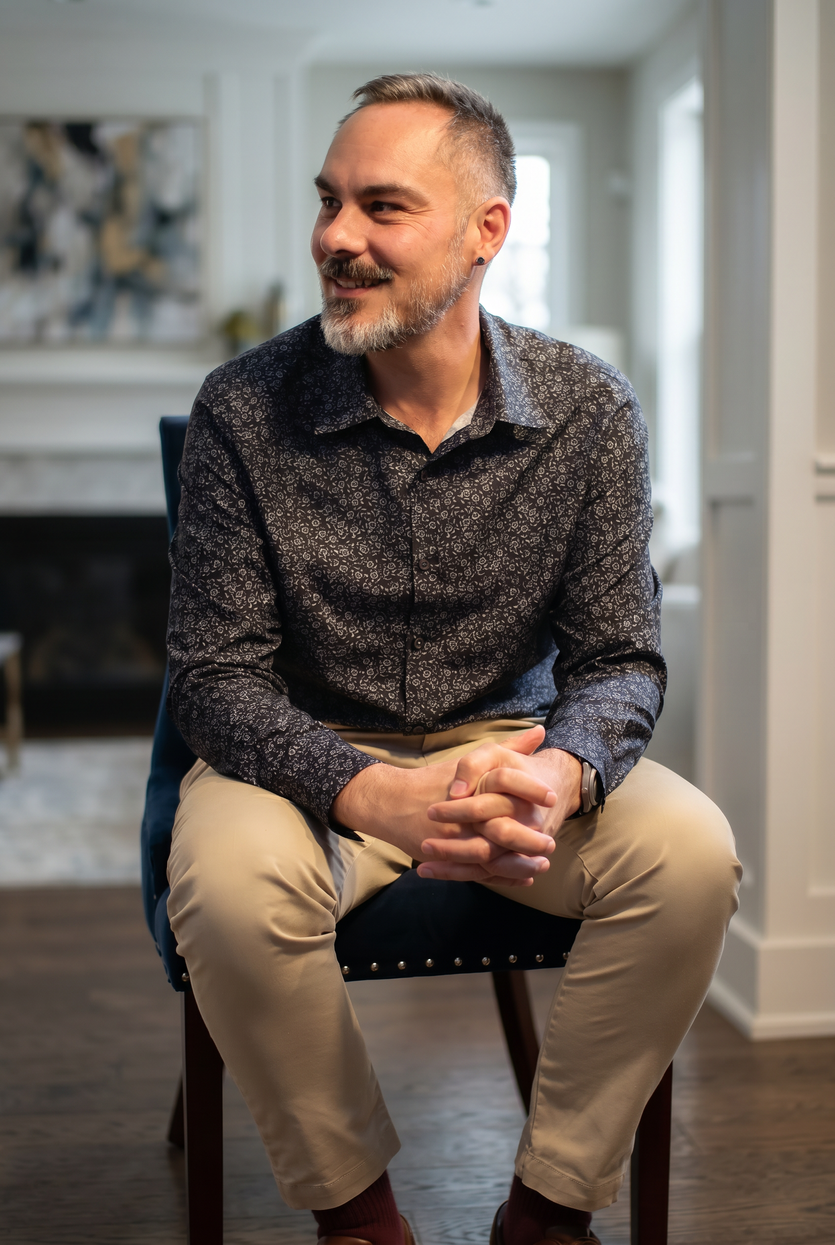 A man sitting on a chair indoors, wearing a dark patterned shirt and beige pants, smiling and looking to the side.