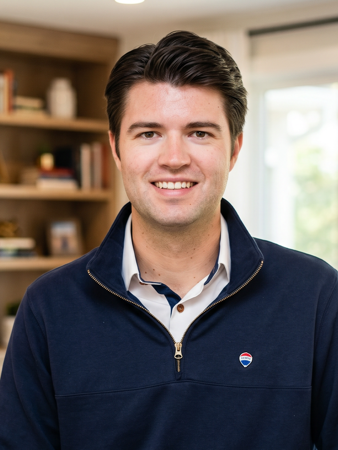 A young man with dark hair smiling in a room with bookshelves and a window in the background.