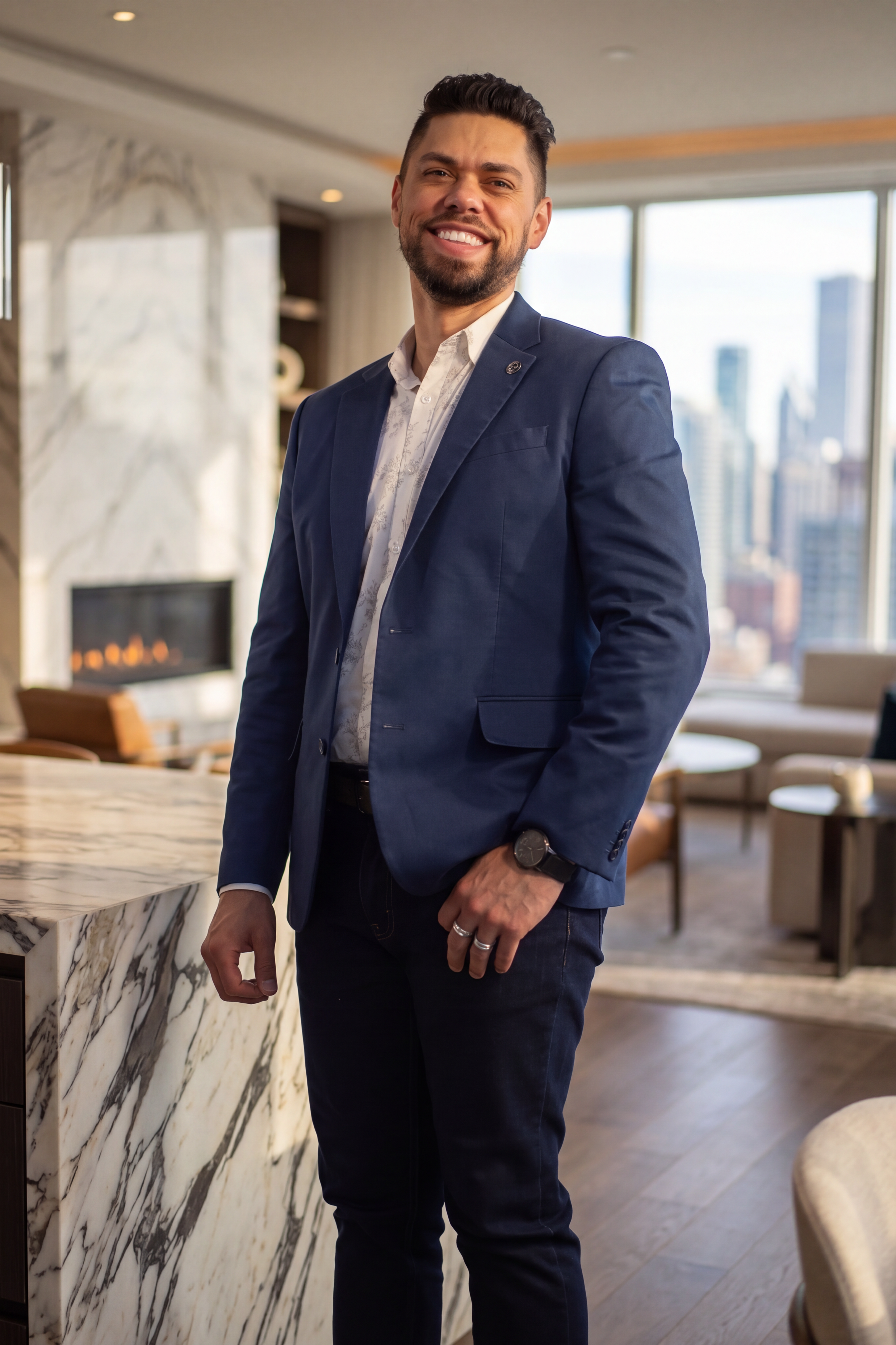 A man in a blue blazer and white shirt smiling in a modern apartment with large windows and city views.