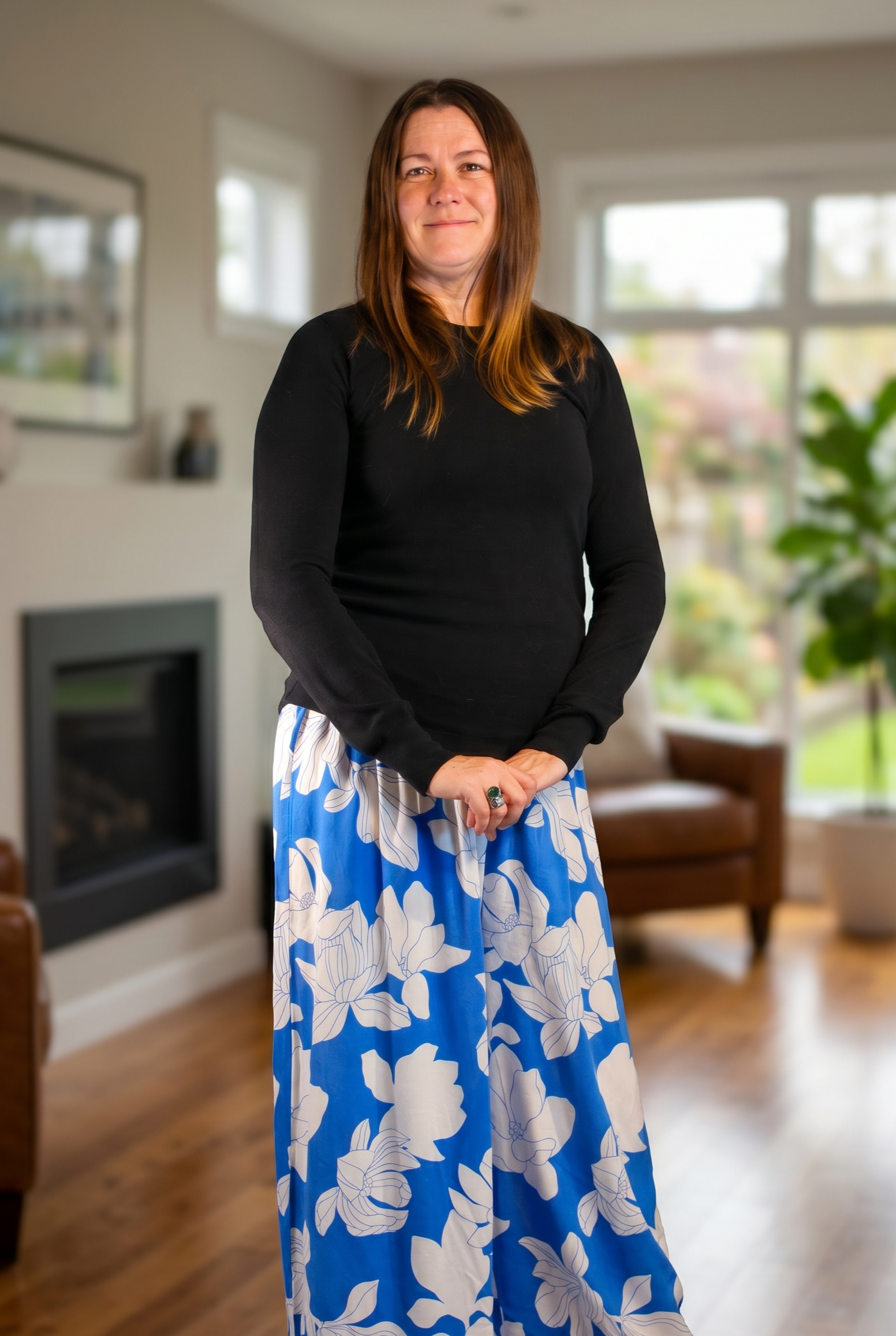 A woman with brown hair wearing a black long-sleeve shirt and a blue and white floral maxi skirt standing in a living room with large windows and wooden flooring.