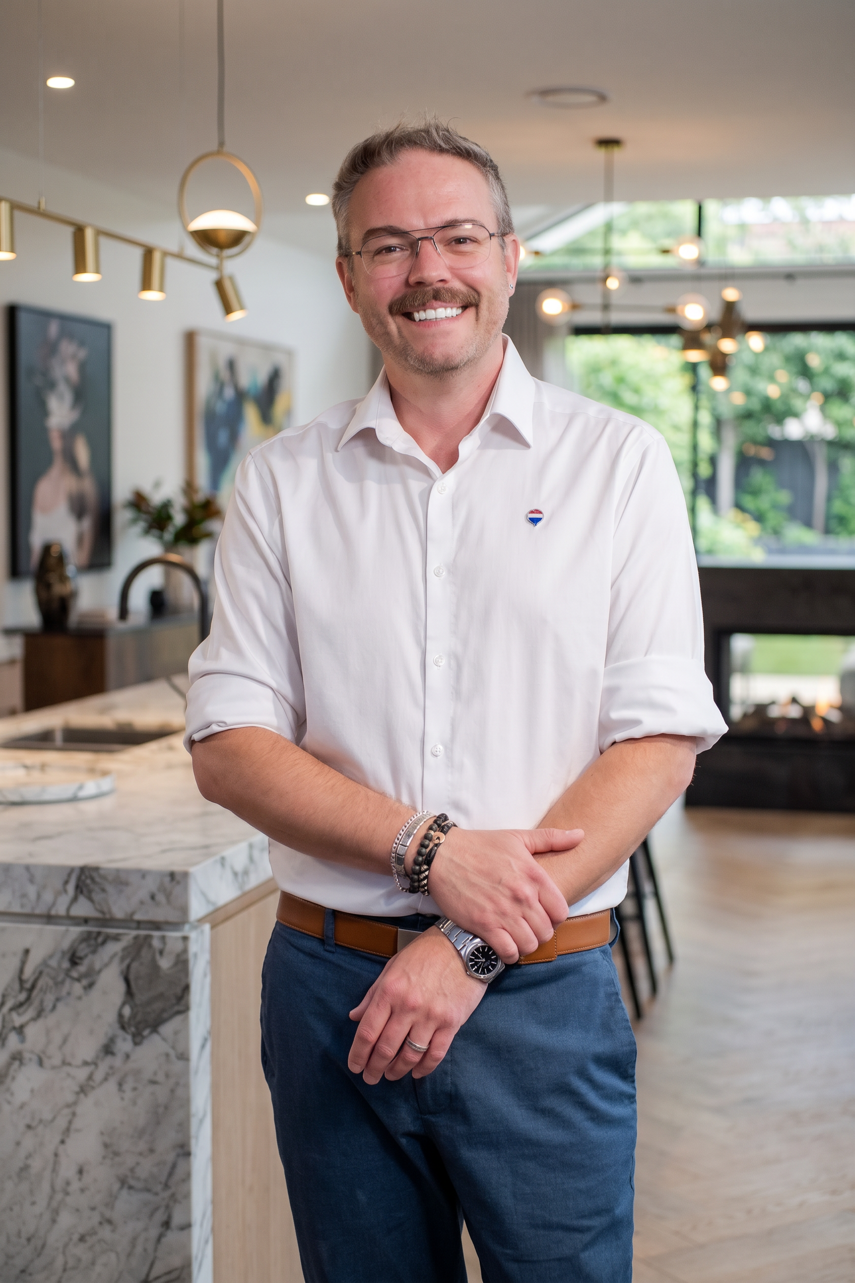 A smiling man with glasses, a mustache, and a beard, wearing a white shirt and blue pants, standing in a modern kitchen with artwork on the wall and a large window with greenery outside.