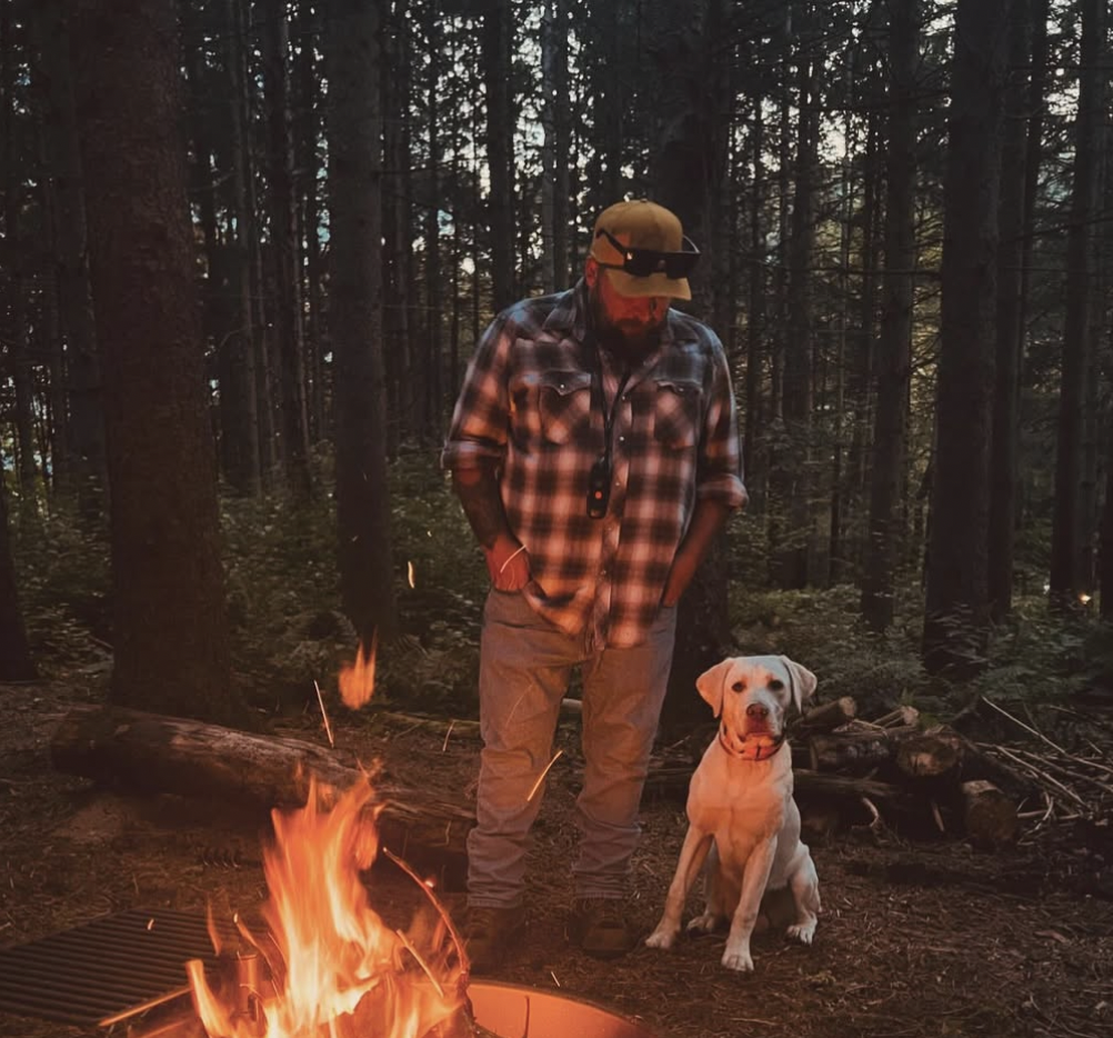 A man in a plaid shirt, baseball cap, and sunglasses standing in a forest with a Labrador retriever sitting beside him near a campfire.