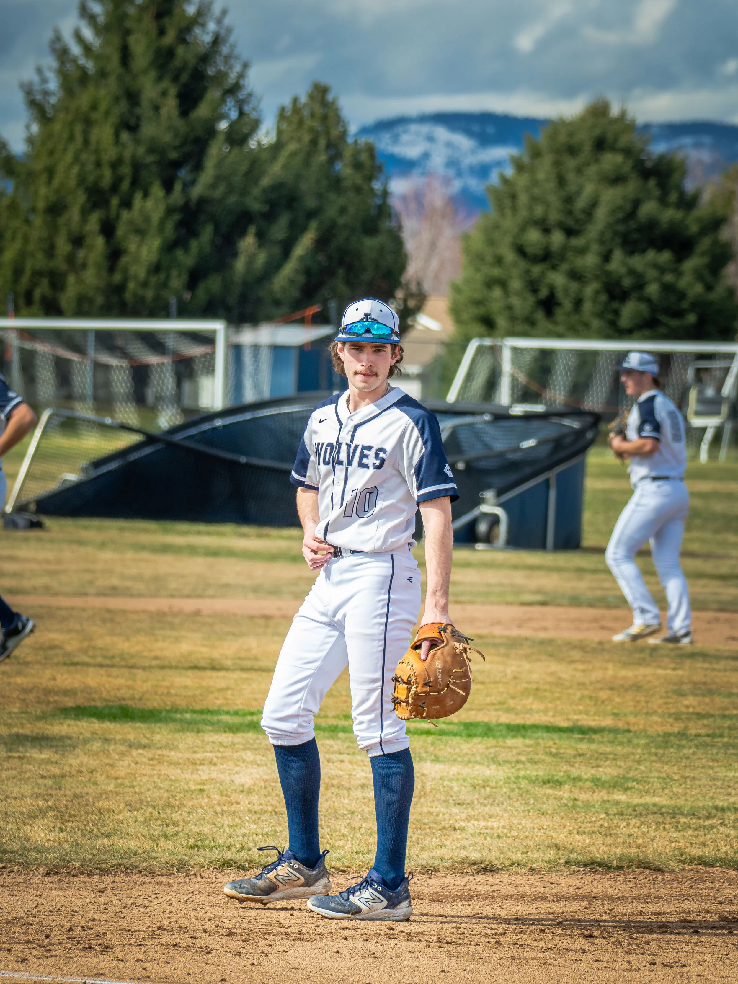 Baseball player in "Wolves" uniform holding glove while standing on field, with trees and mountains in background.
