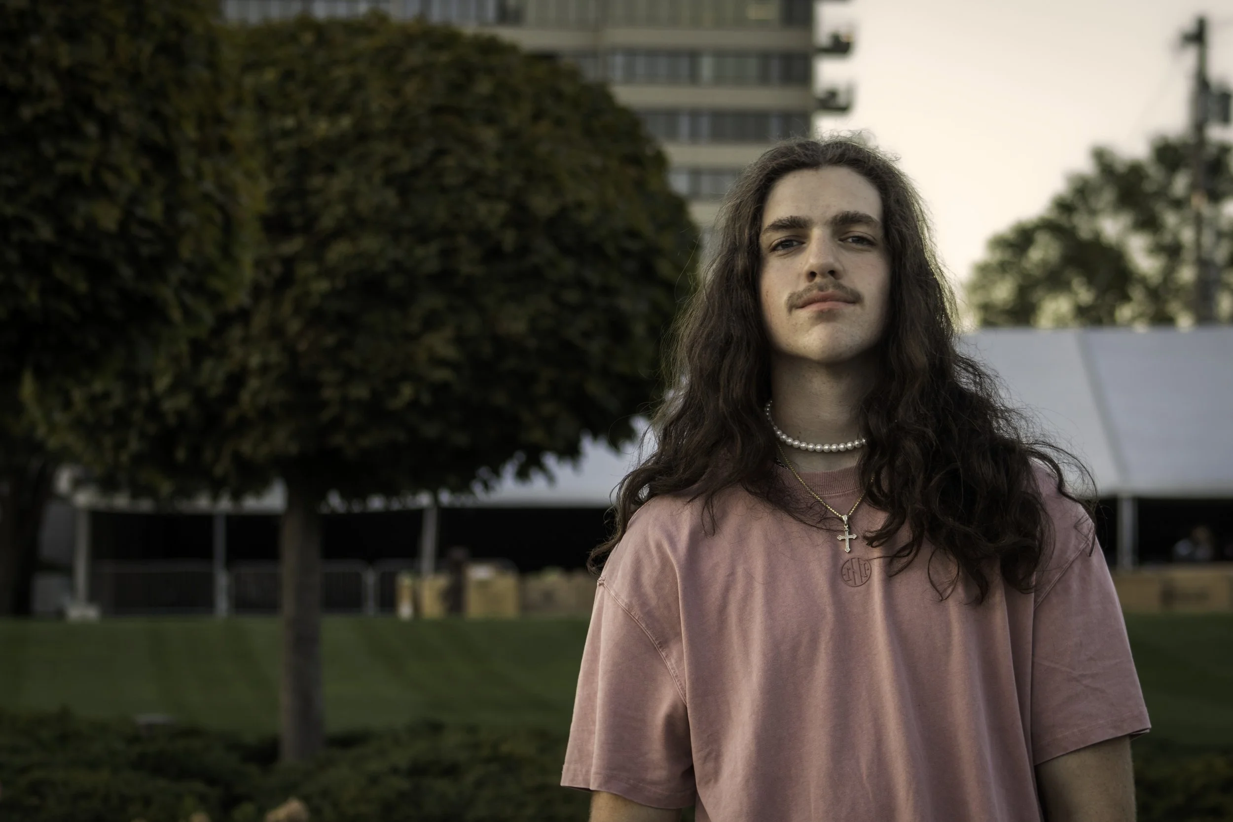 A young person with long, curly brown hair and light skin standing outside in a park-like setting, wearing a pink t-shirt, pearl necklace, and a gold chain with a cross pendant, with trees and a building in the background.