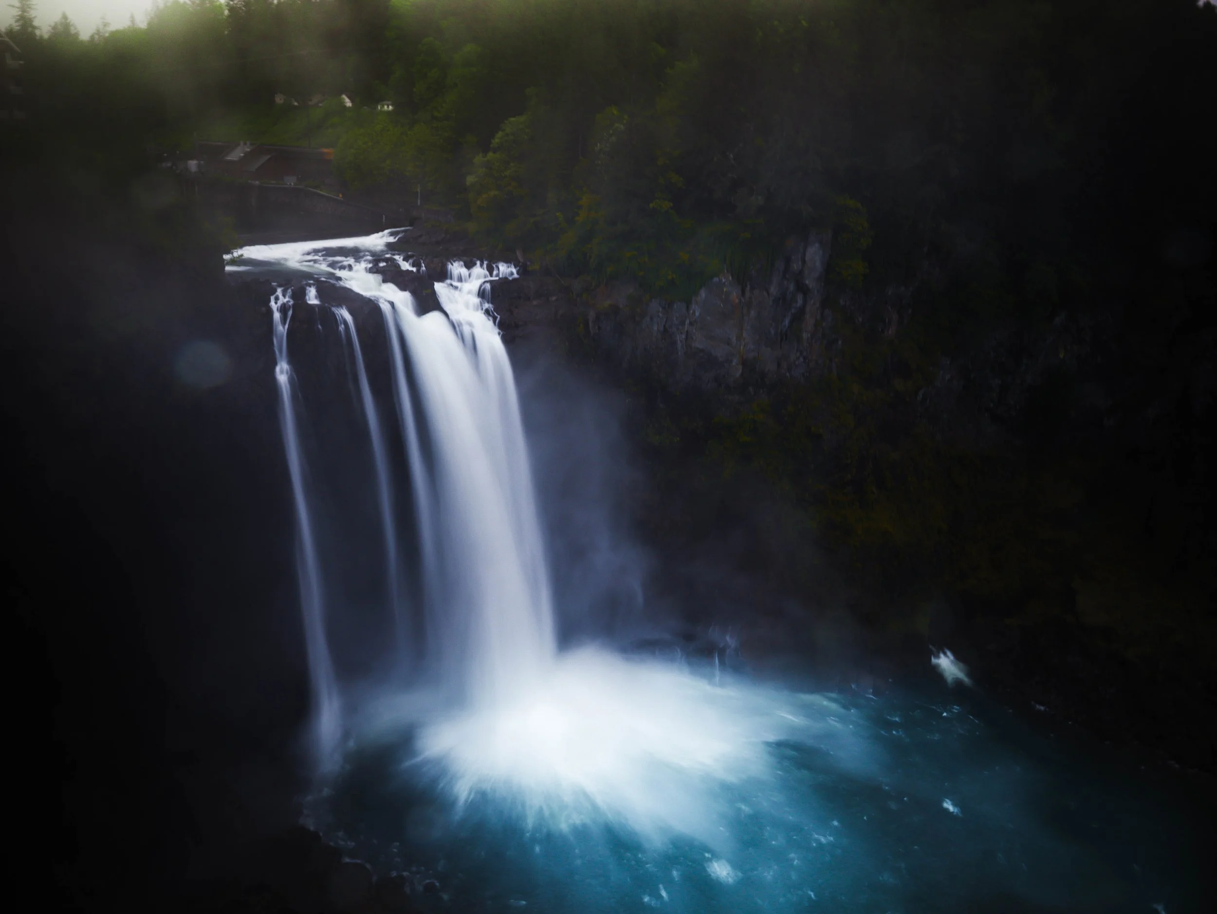 A waterfall cascading into a pool surrounded by dark rocks and lush greenery, with trees in the background.