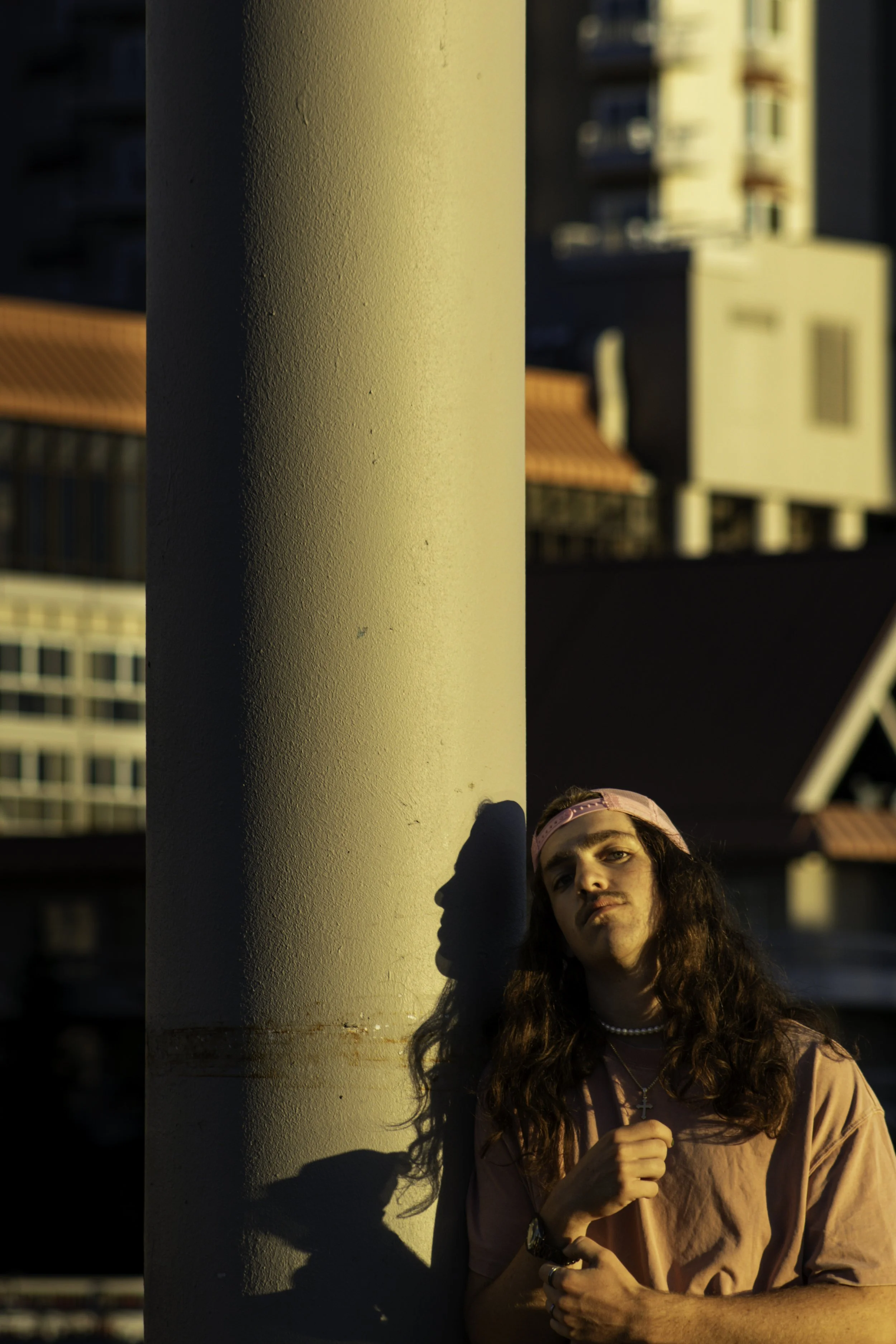 Person with long hair, wearing a pink shirt and cap, leaning against a concrete pillar at sunset, creating a shadow on the pillar.