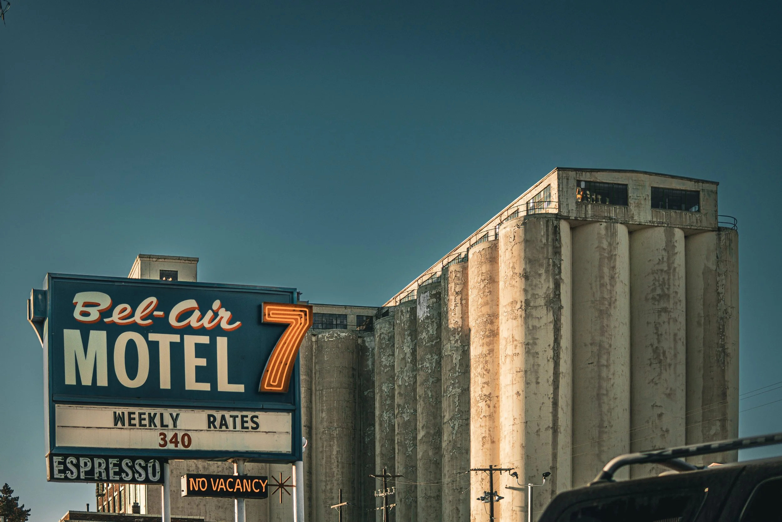 Vintage motel sign reading "Bel-Air Motel 7" with weekly rates and no vacancy, set against an old industrial building with silos, under a clear blue sky.