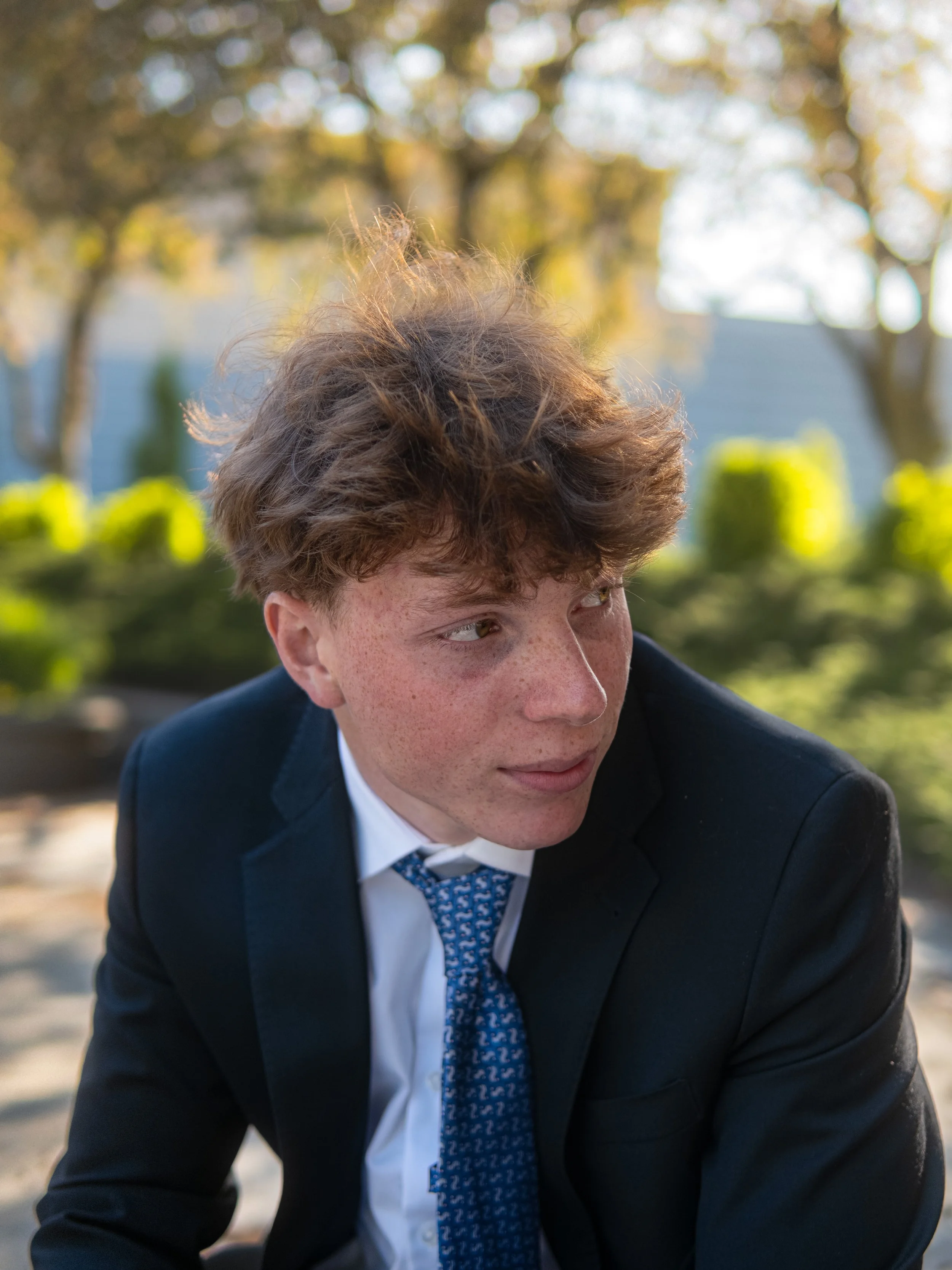 A young man with reddish hair, freckles, and dressed in a black suit with a white shirt and blue patterned tie, is sitting outdoors. The background is blurred with sunlight filtering through trees and greenery.