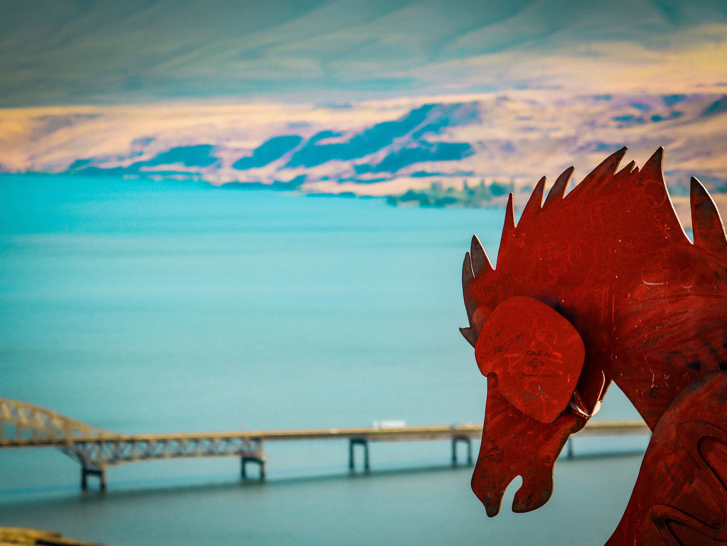 A rusty metal sculpture of a fish near a body of water. In the background, a bridge structure extends over the water with a cloudy sky above.