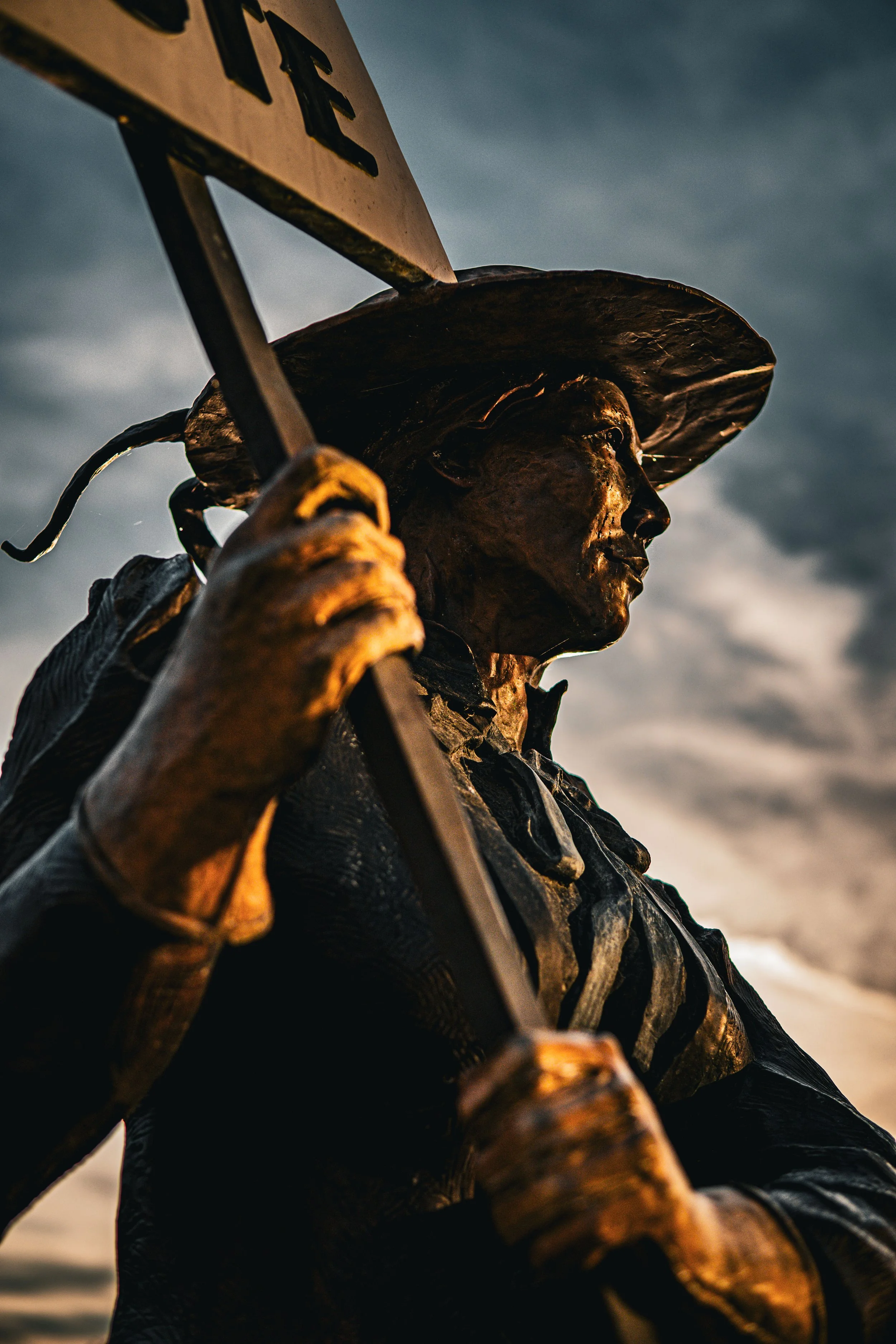 Bronze statue of a person holding a sign, wearing a wide-brimmed hat, against a cloudy sky background.